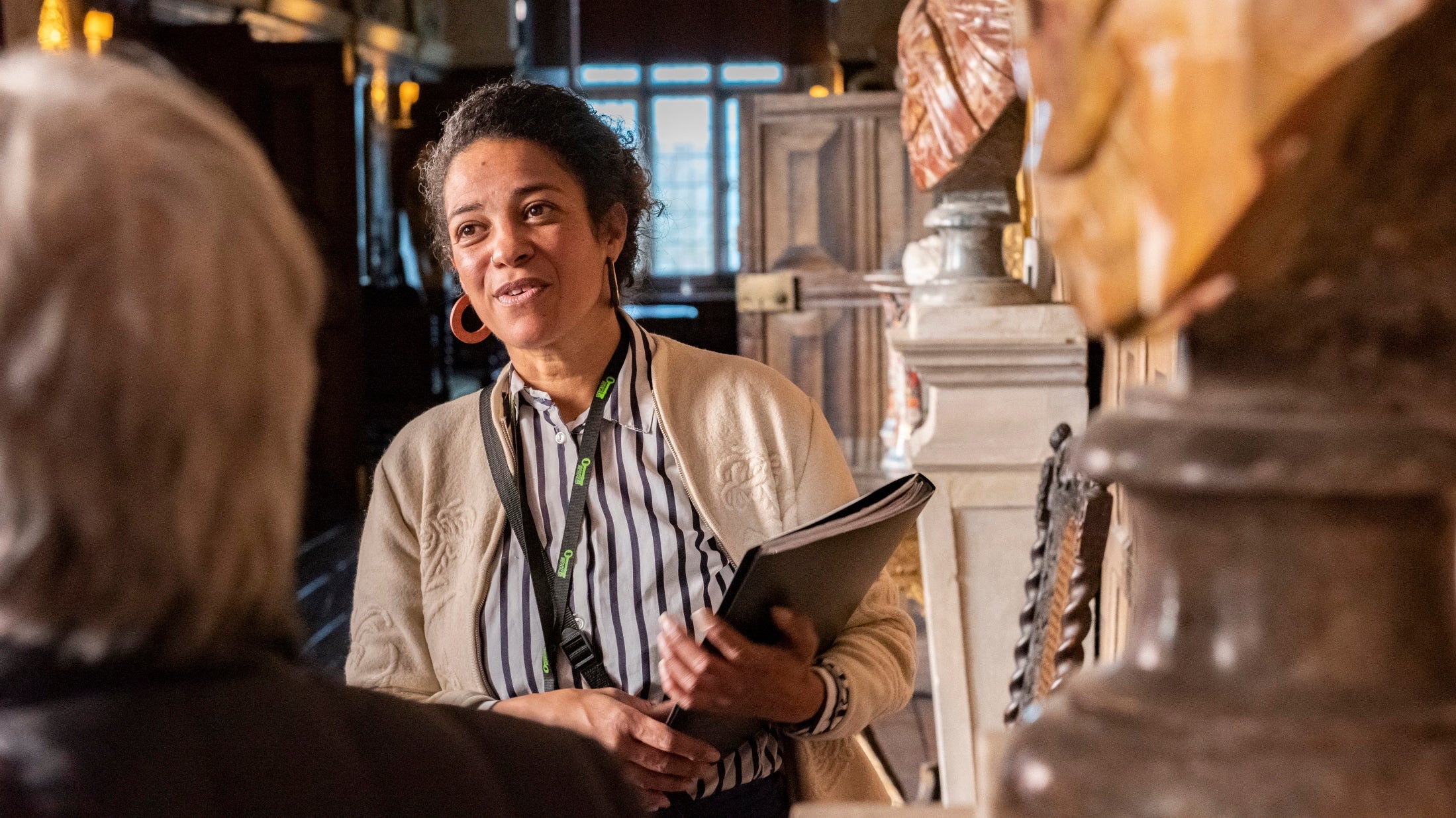 A woman wearing a black and white striped blouse, carrying a folder is mid-sentence talking, possibly about the wooden carved statue next to her.