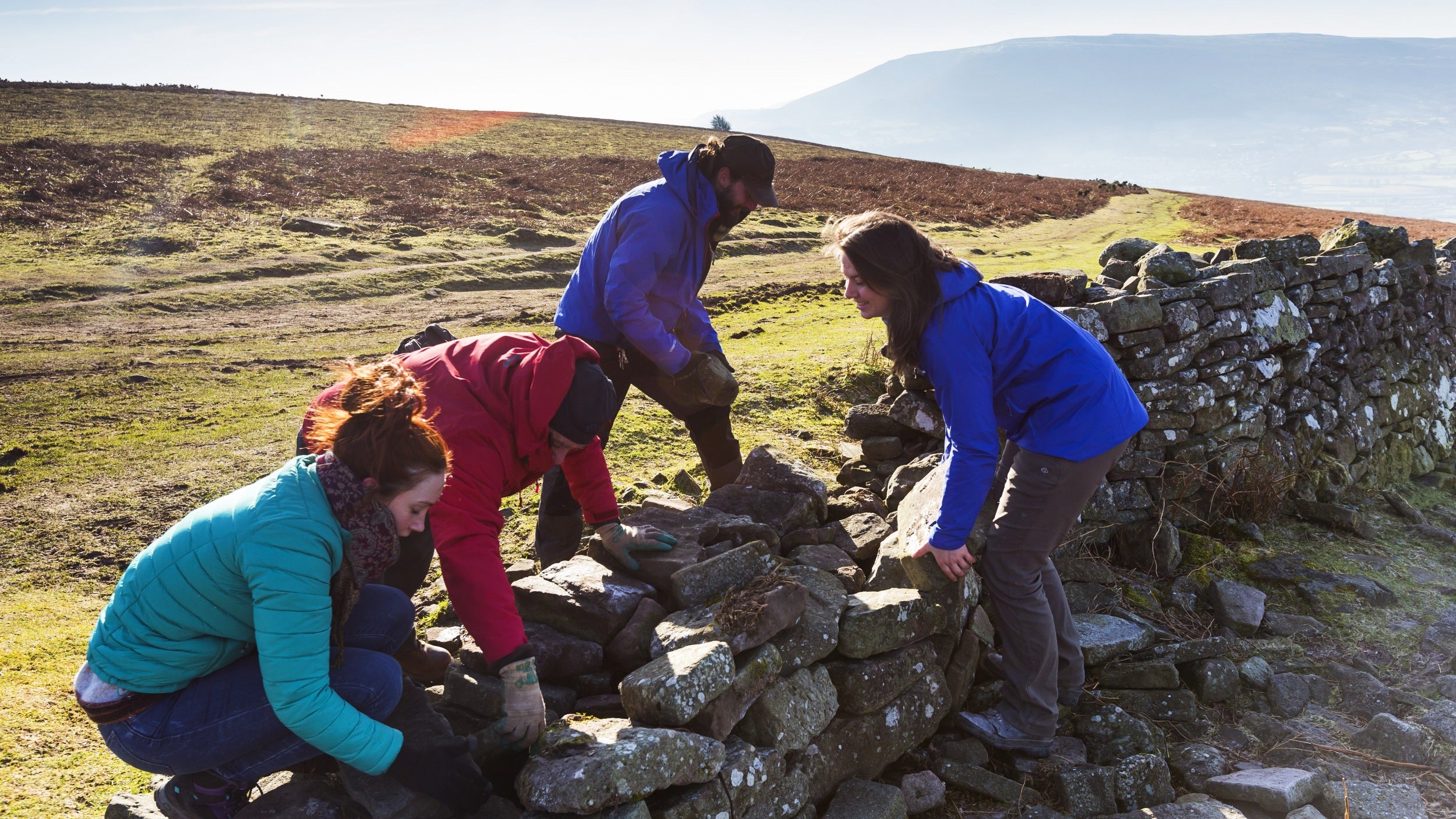 A group of four twenty-somethings wearing brightly coloured outdoor jacked are lifting and placing large stones on a broken wall in a landscape of rolling green hills.