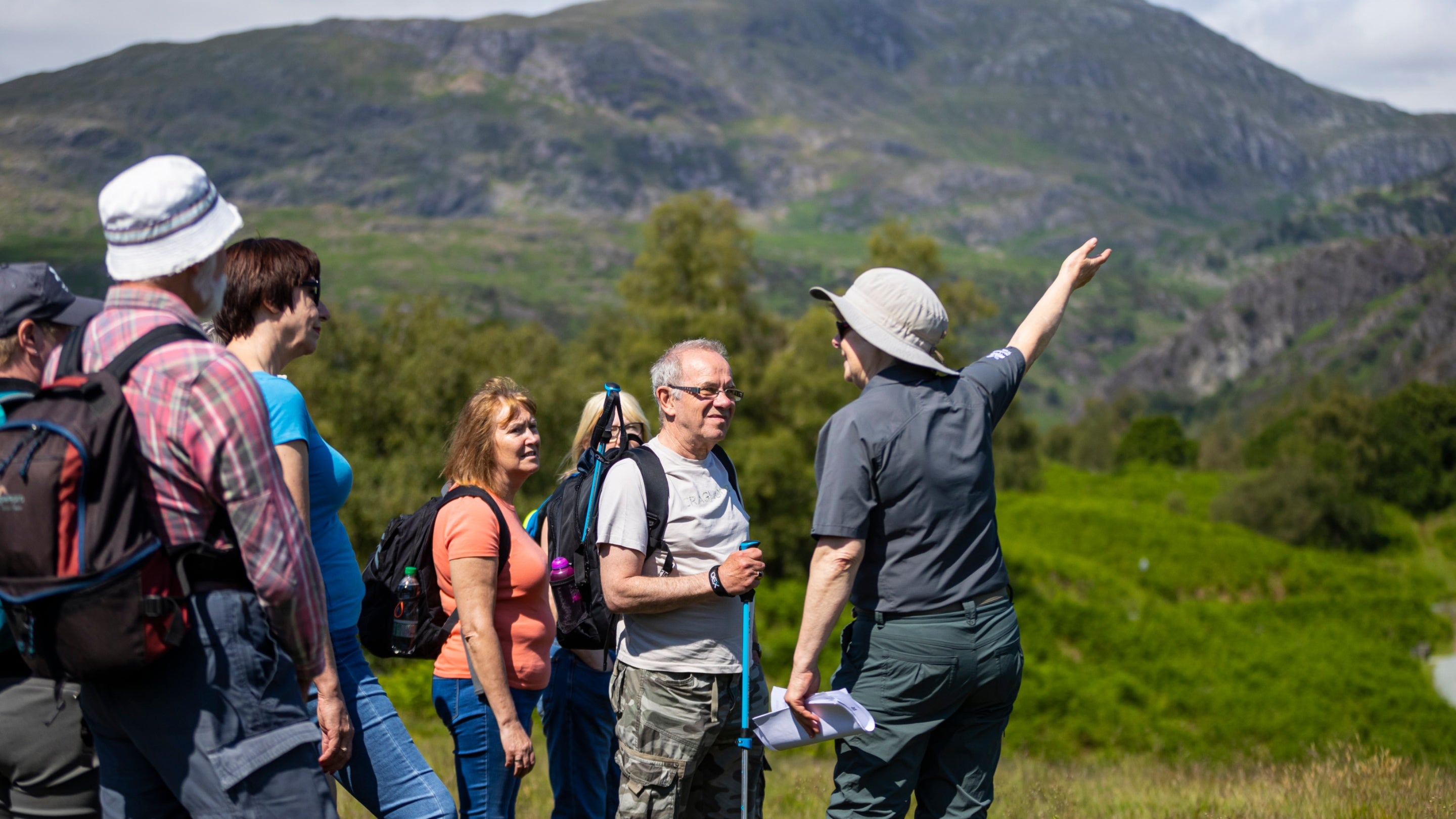 Image shows a group of people on a guided walk with a volunteer at Tarn Hows on a sunny day, with a view of a hill in the background