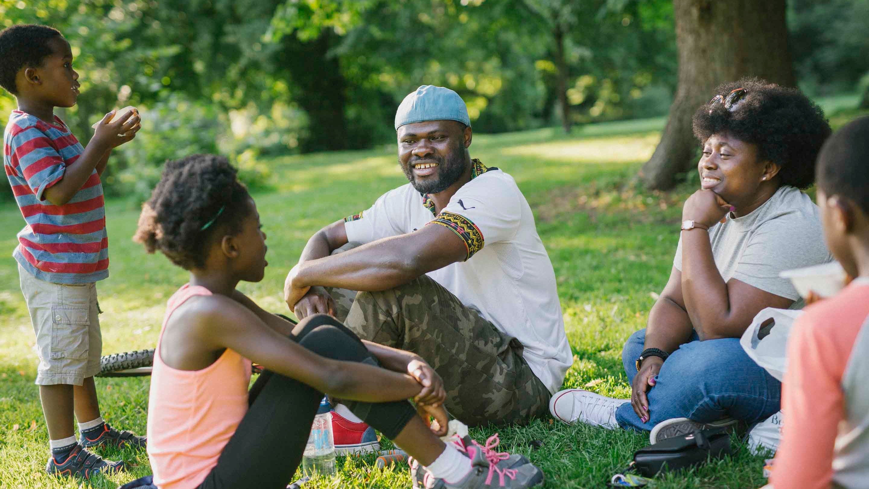 A family group of five people sit on the grass chatting at Morden Hall Park in the summer. Patches of sunshine break through the mature tree canopy.