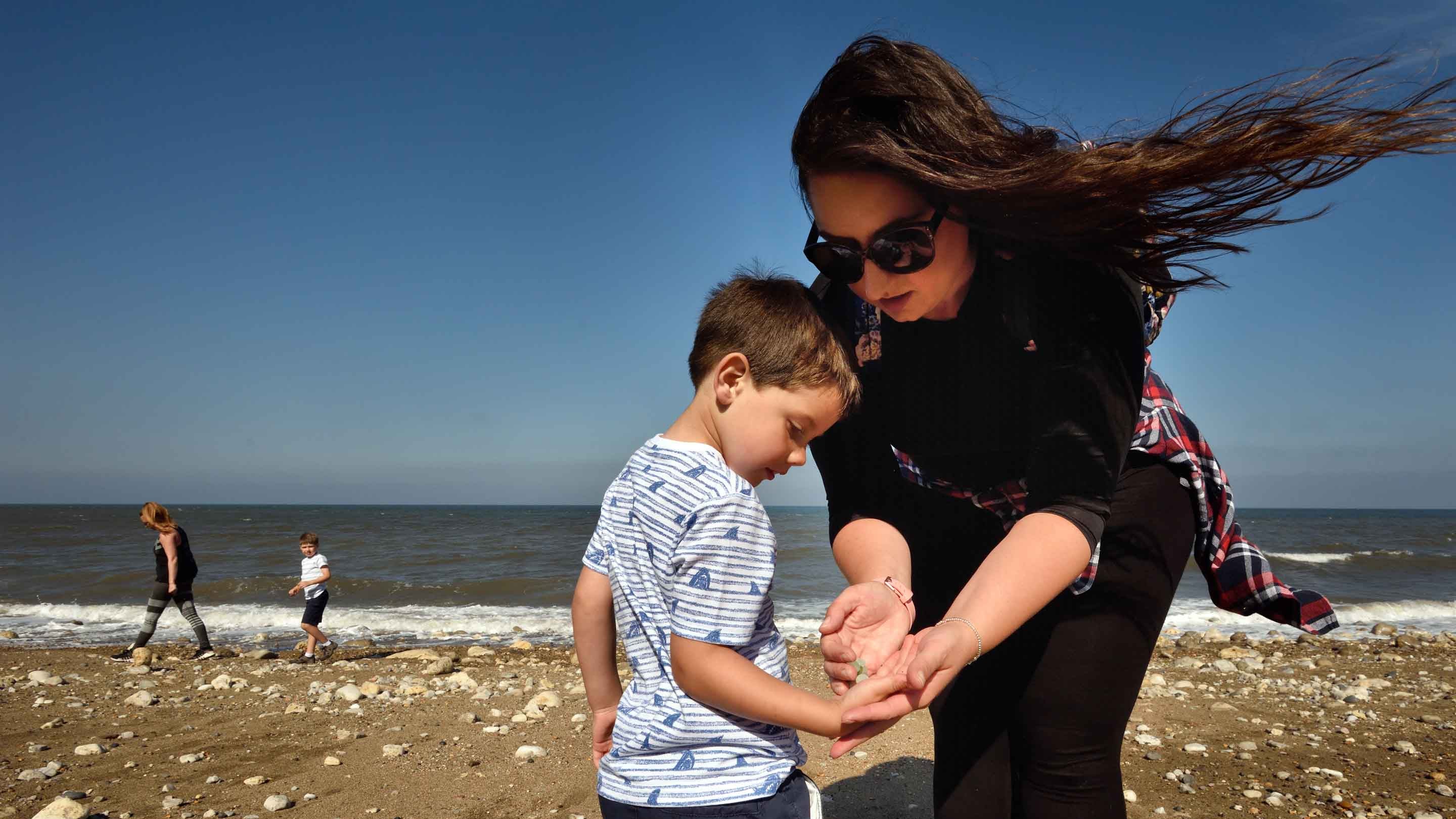 A woman and boy look at sea treasure in their hands whilst two other people walk along the tideline in the background