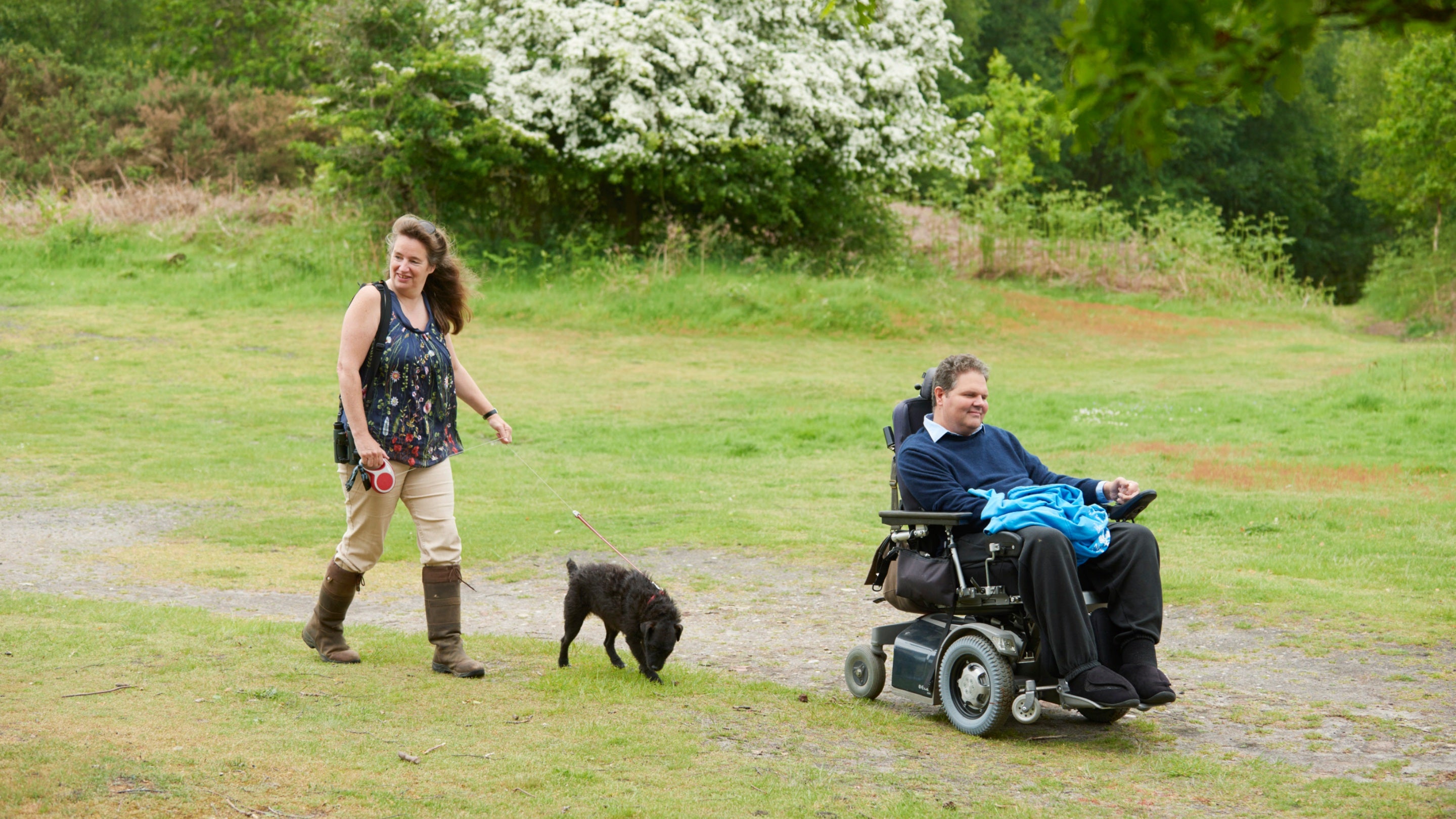Man in electric wheelchair and woman walking dog using gravel path through field