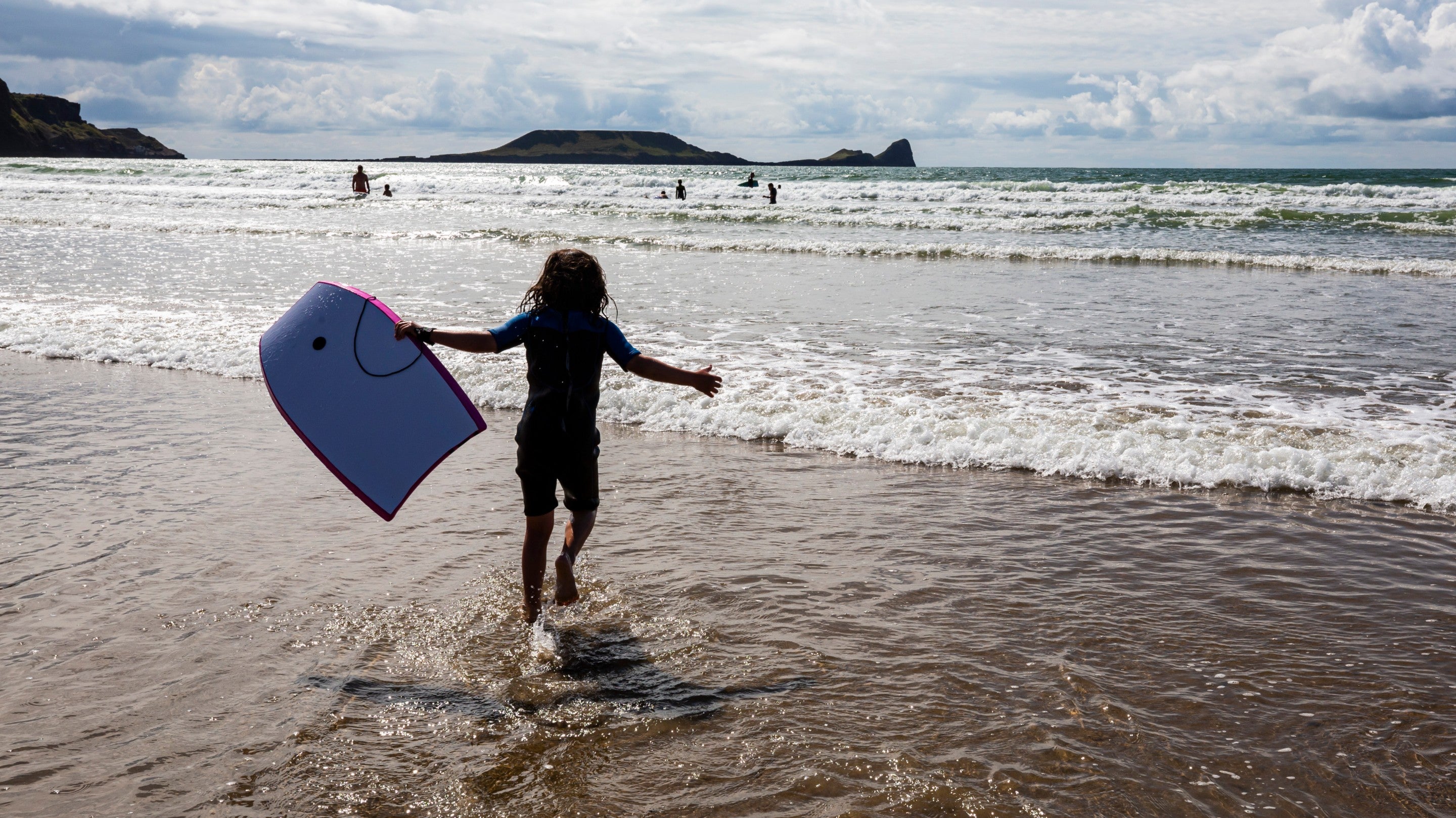 The silhouette of a child running into the sea with a bodyboard at Rhosili and South Gower Coast, Wales