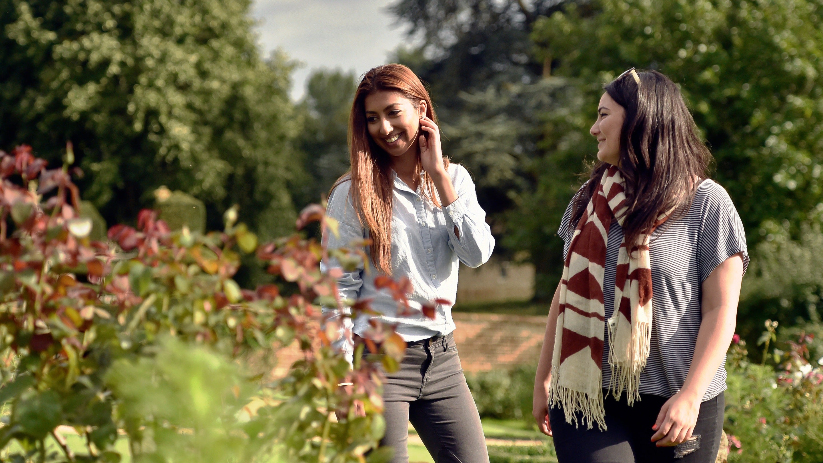 Two visitors walk in the gardens at Canons Ashby, Northamptonshire. Rose bushes can be seen in the foreground with mature trees in parkland behind.