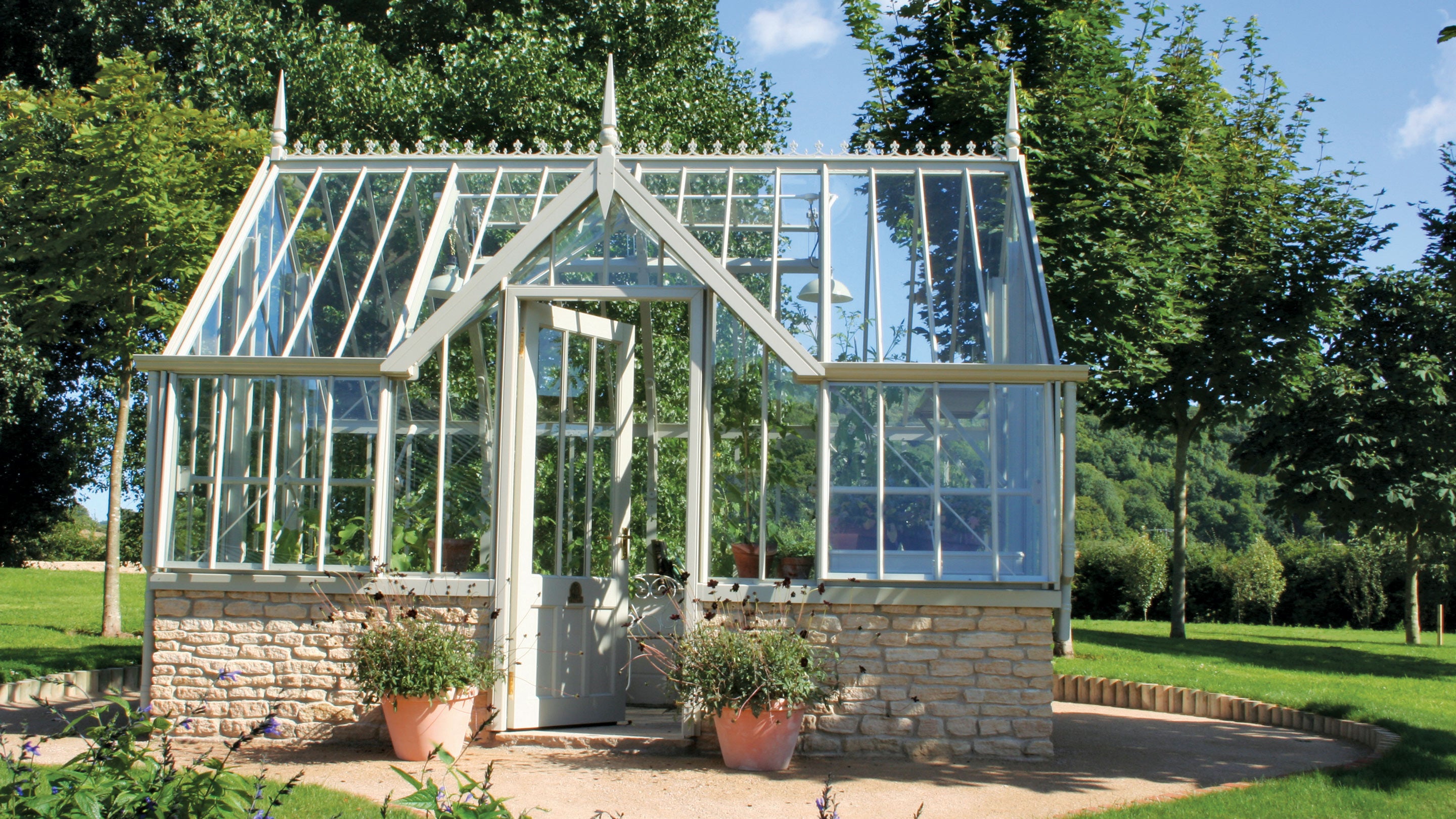 A Victorian style glasshouse sits on a small patio surrounded by lawn and established trees.