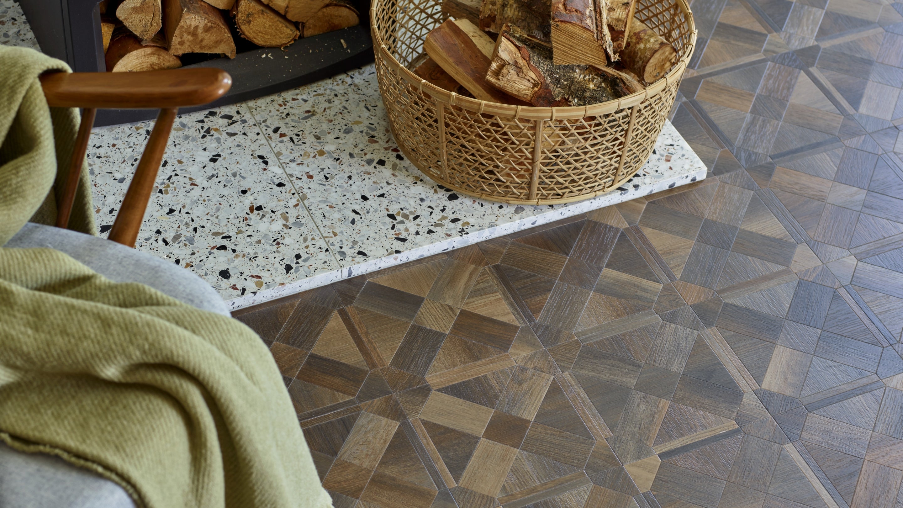A patterned timber floor surrounds a stone fireplace with a log basket, chair and blanket nearby