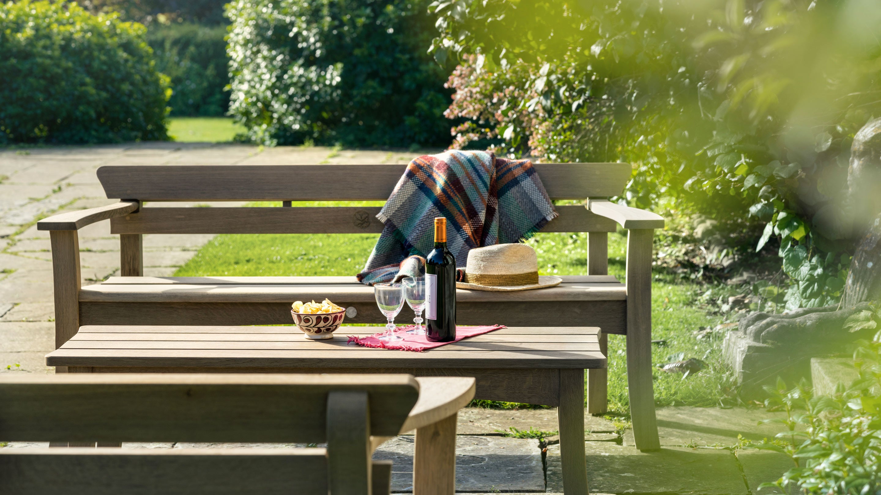 Low shot of a wooden bench and table on a stone patio surrounded by shrubbery