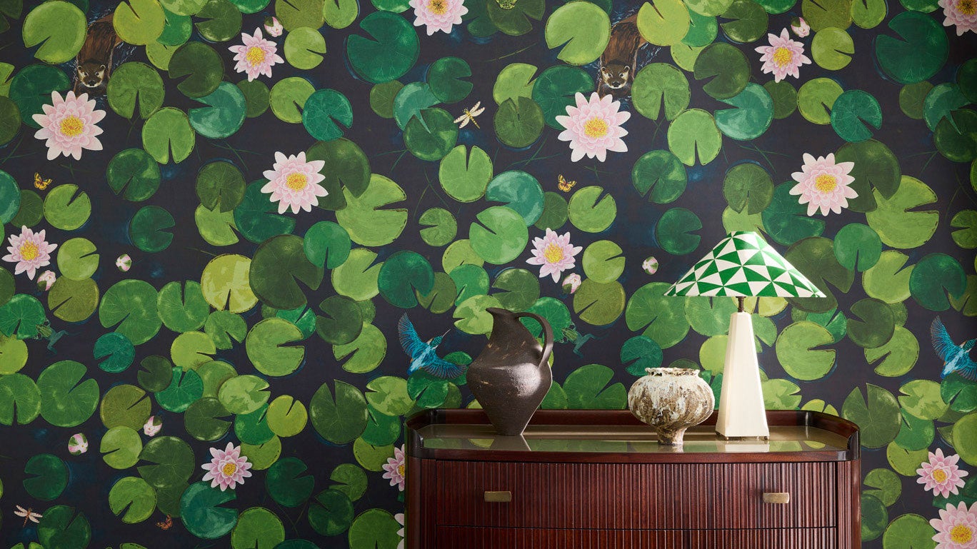 Wallpaper with a bold water lilly print in deep greens and pink sits behind a sideboard with a lamp and vases on top.
