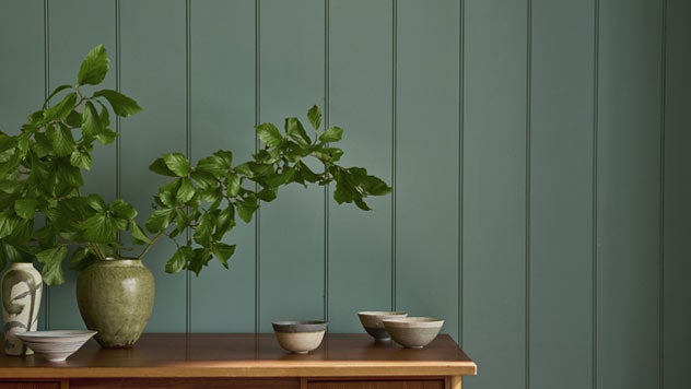 A wooden side table sits in front of a tongue and groove wall painted in a grey green.