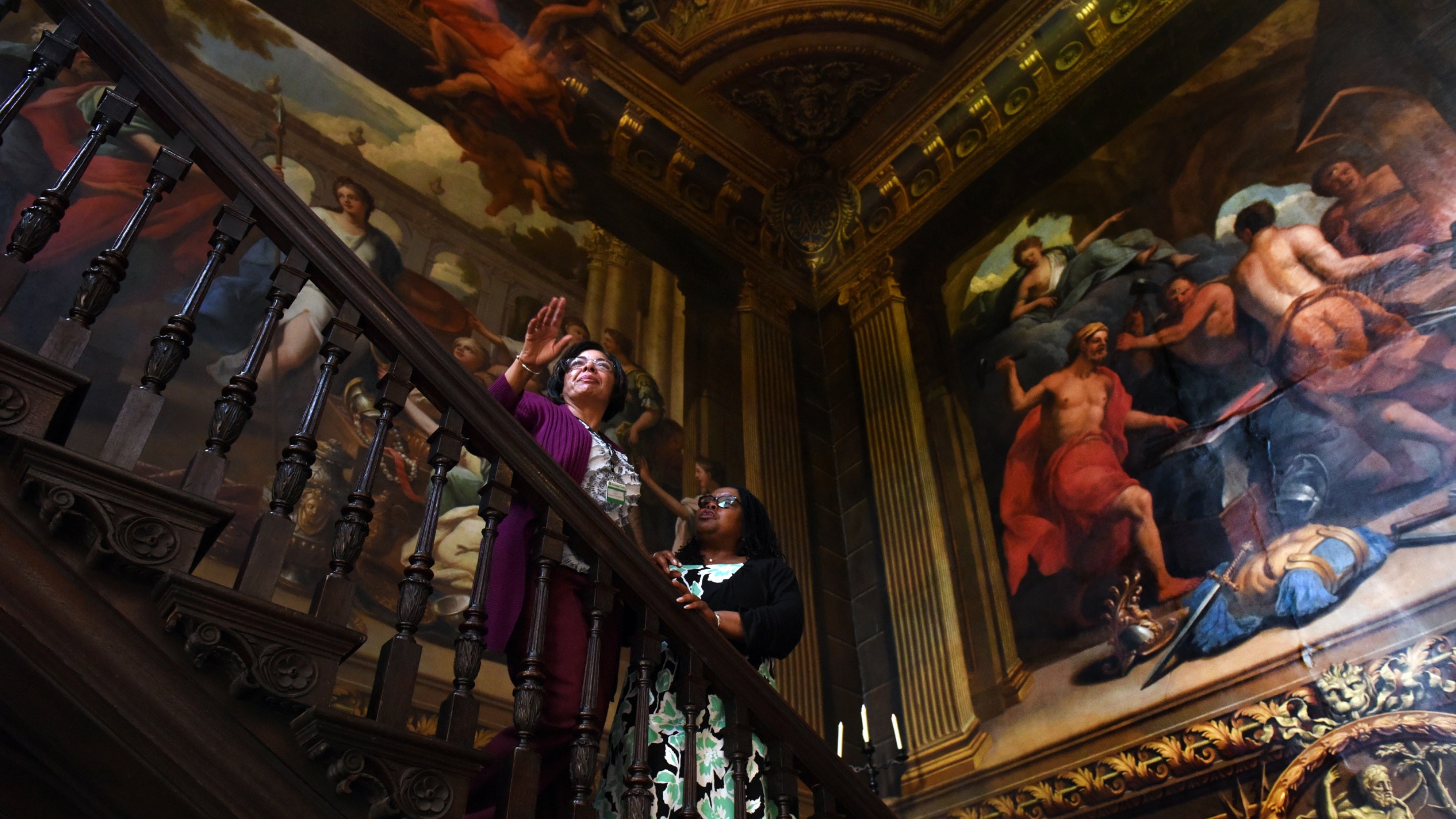 Two visitors standing on an ornate dark wood staircase surrounded by large colourful classical paintings covering the walls