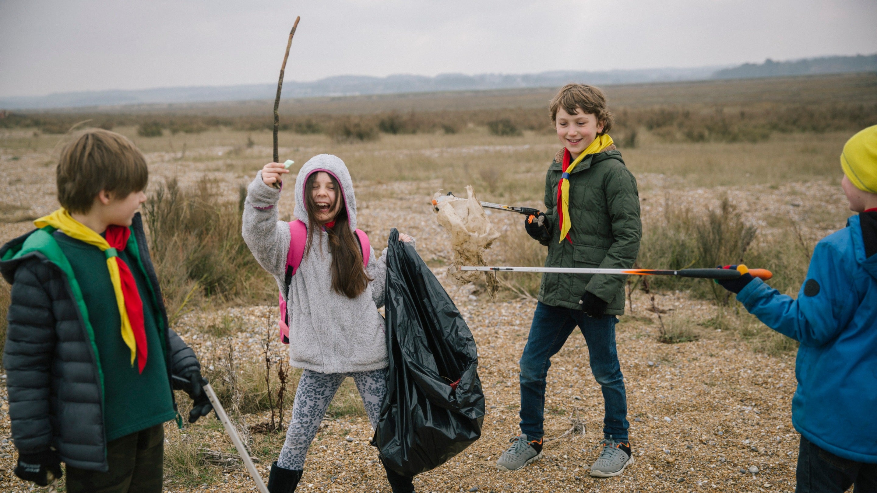 Four children, two dressed in Scouts uniforms, posing on a beach with equipment for rubbish picking