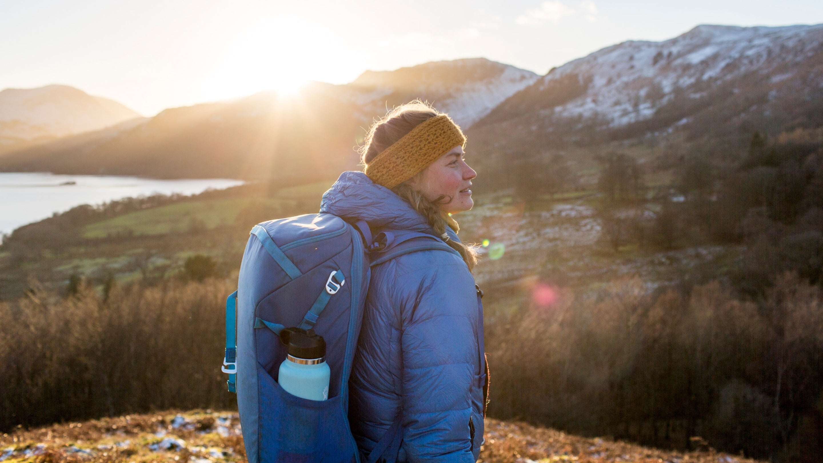 A young woman in winter clothing looking over a landscape of hills with the sun setting