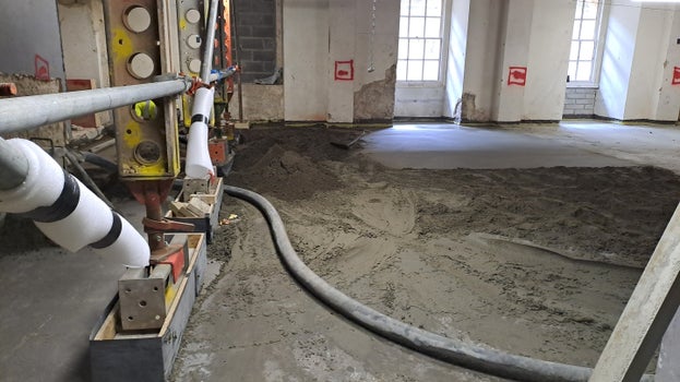 A pipe is led across the floor pumping in the loose screed, patches can be seen that have already been smoothed and are drying .
