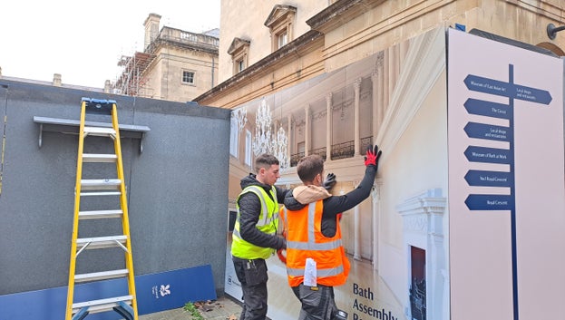 Two people are holding graphics panels up against the hoarding, one is holding a nail gun to hold the graphics in place. Bath Assembly Rooms can be seen behind the hoarding.