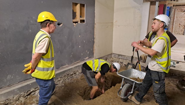 A group of people can be seen working in the basement. One is digging a trench and lifting the soil into a wheelbarrow whilst being observed by others.