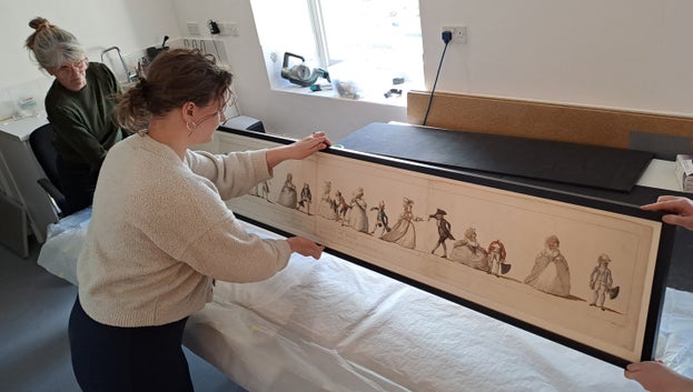 Three people are lifting the framed Long Minuet print from the tissue paper that is led on the table below.
