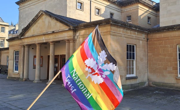 A Pride flag is being waved outside the entrance to Bath Assembly Rooms, the building can be seen behind the flag.