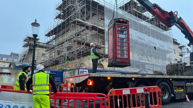 The K6 telephone box is suspended by a crane above the bed of a lorry being guided down by a person holding controls.  In the background Bath Assembly Rooms can be seen covered in scaffolding.