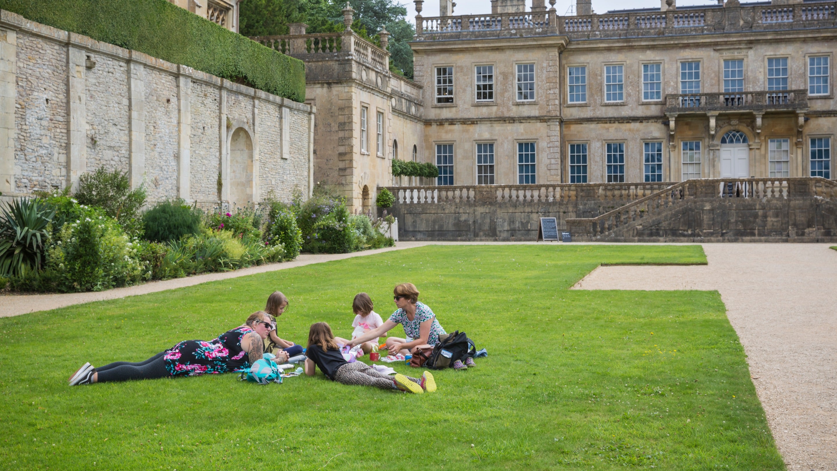 A small group of adults and children sit and lie on the grass enjoying a picnic in the summer with Dyrham house in the background at Dyrham Park, Gloucestershire