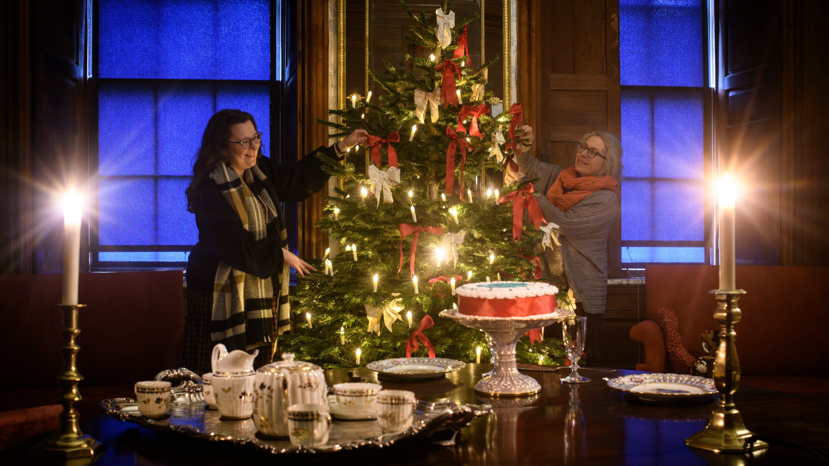 Two members of staff adjusting a Christmas tree behind a table laid out with a tea set and cake