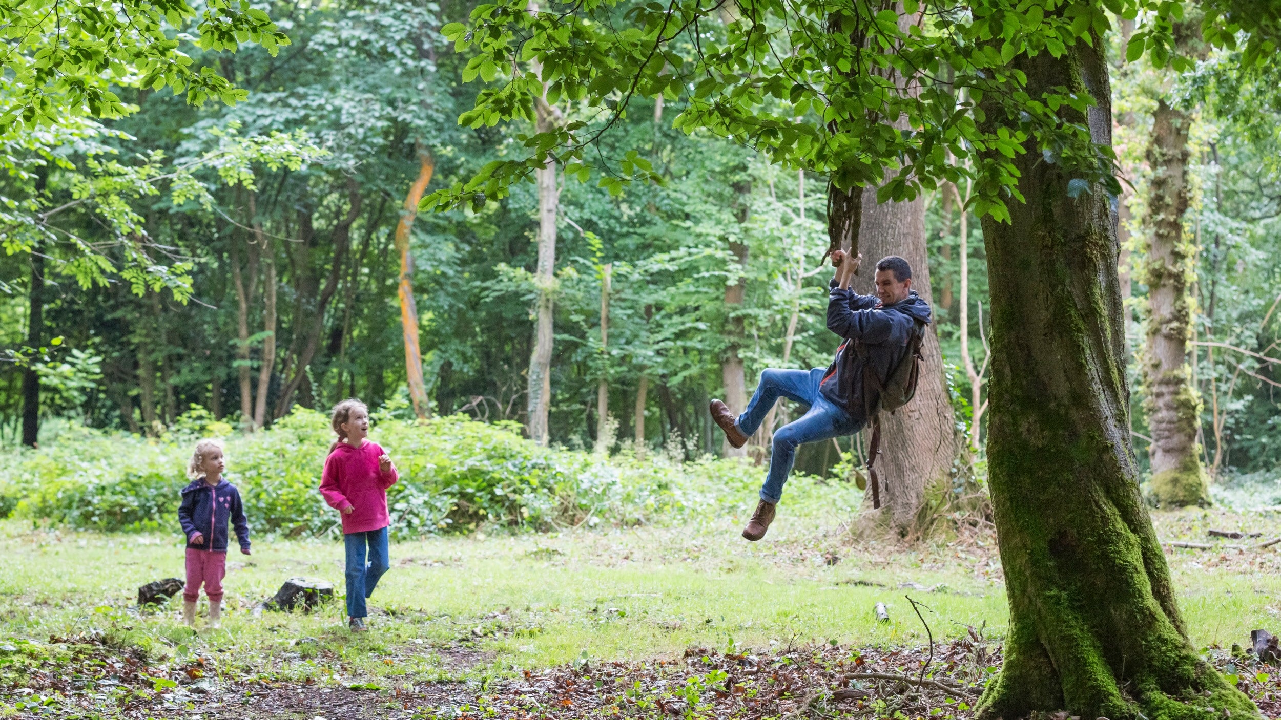 Man swinging on rope swing in woodland while two children look on