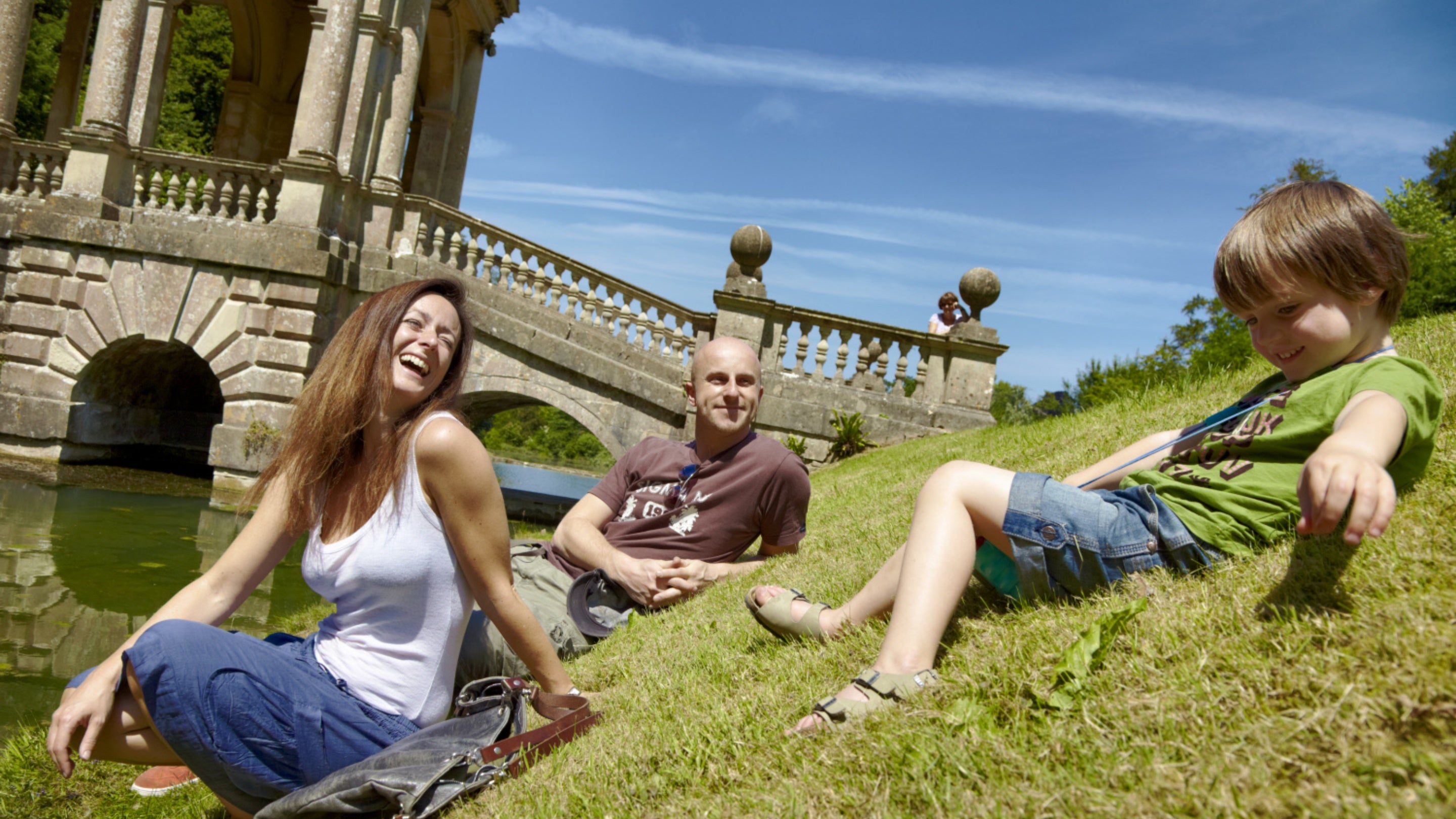 Family visitors on the bank beside the lake with the Palladian Bridge in the background at Prior Park Landscape Garden, Bath