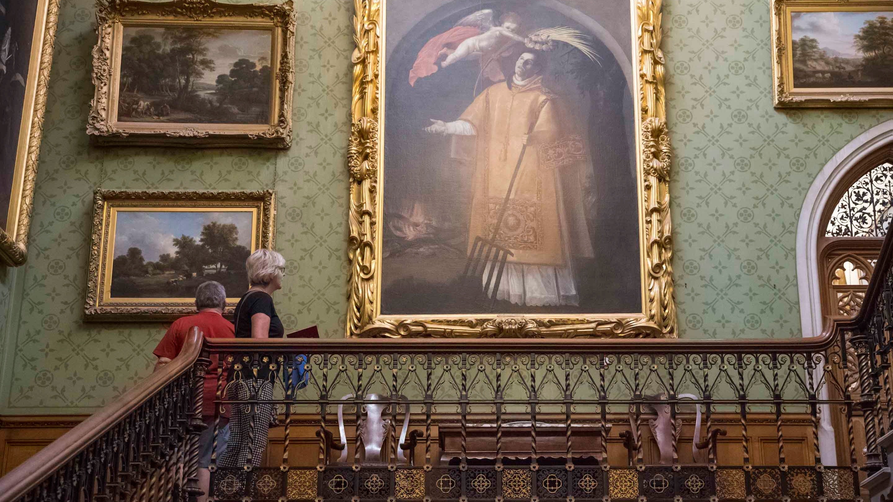 Two people looking at huge portraits and landscape paintings in the Staircase Hall at Tyntesfield