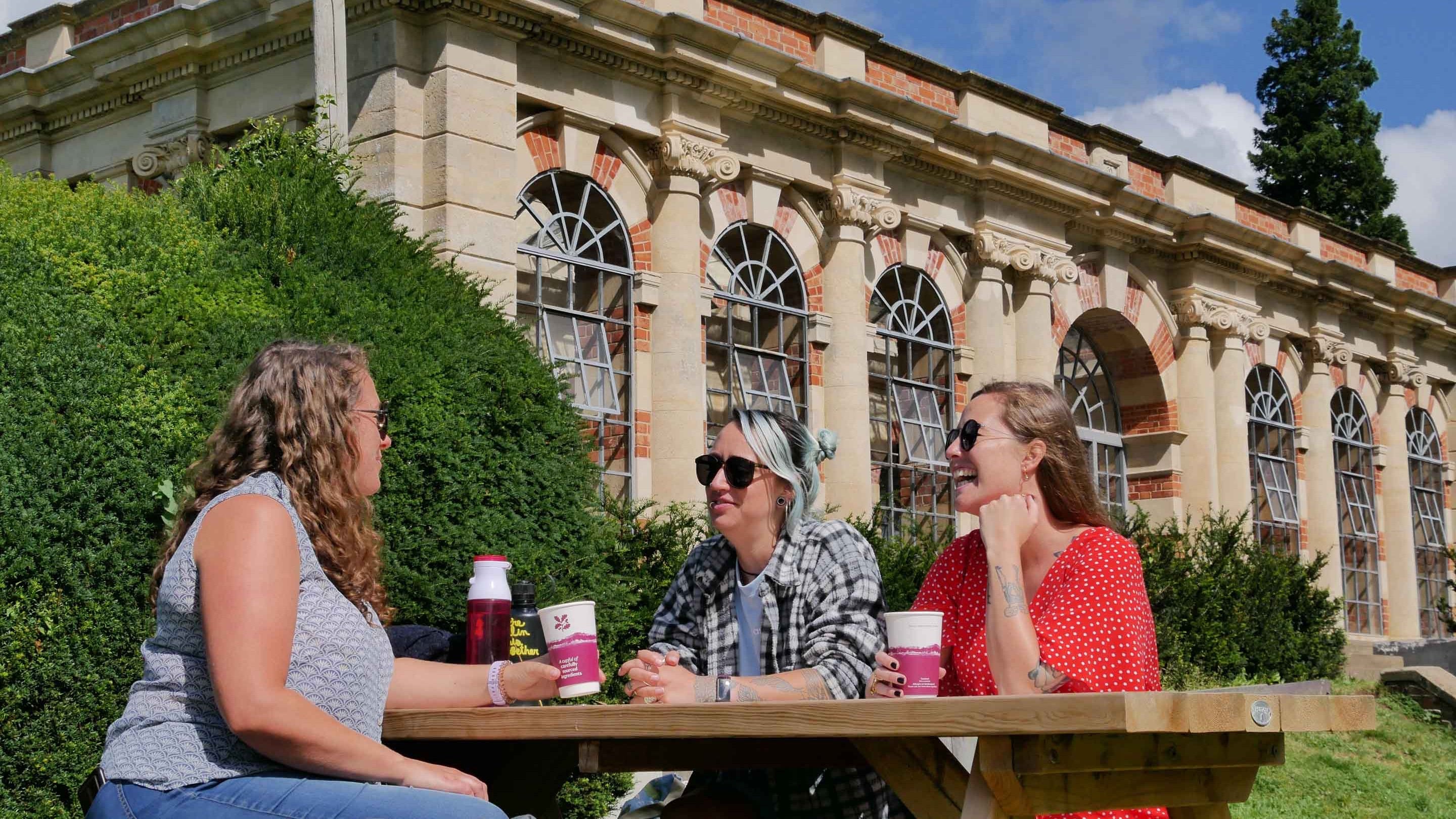 Three visitors enjoy takeaway hot drinks sat on a bench in the garden at Tyntesfield, Somerset