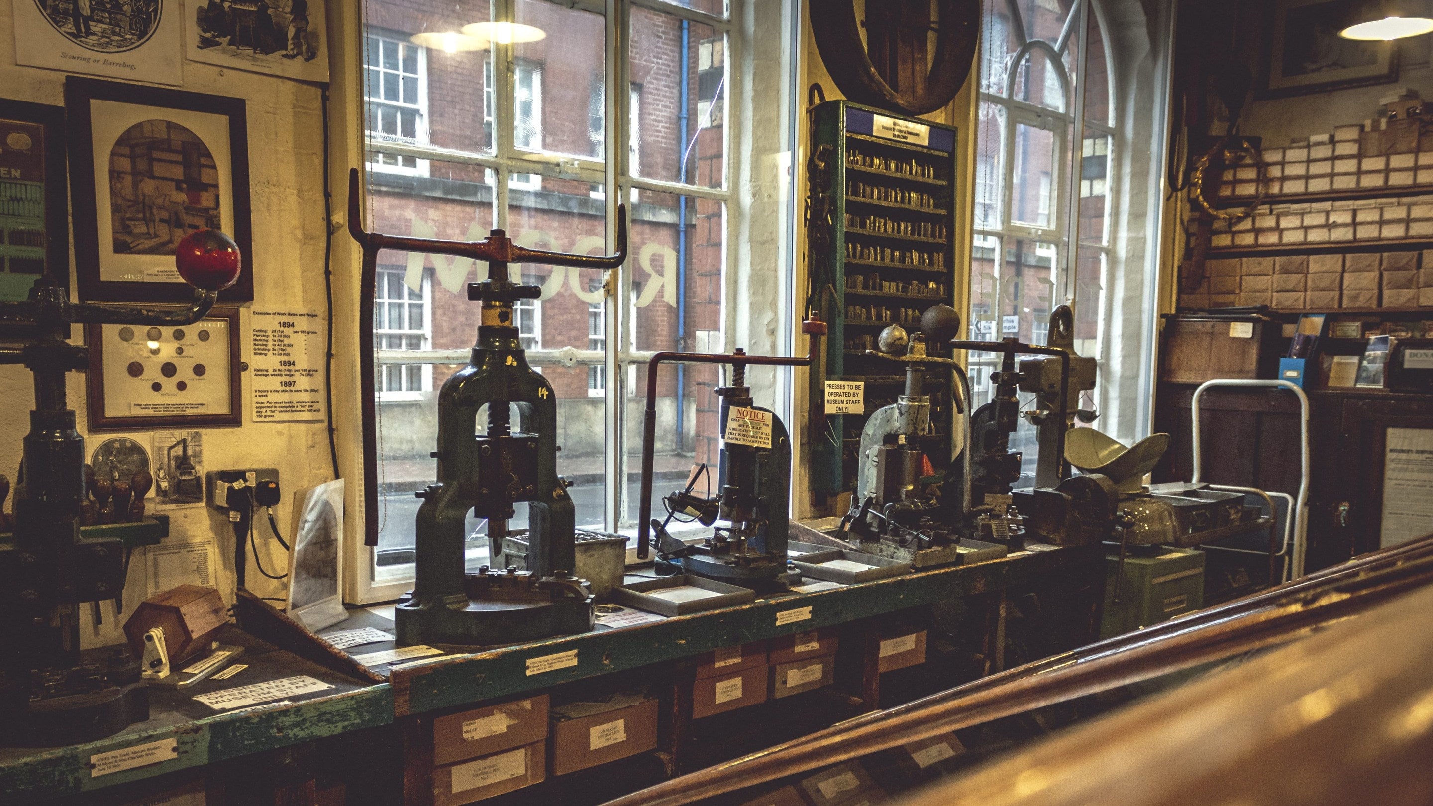 A selection of upside-down U shaped presses line an old wooden bench along a window sill. Beyond the windows are red-bricked buildings and inside the room with the presses are lots of small boxes and wooden Victorian looking items or equipment.