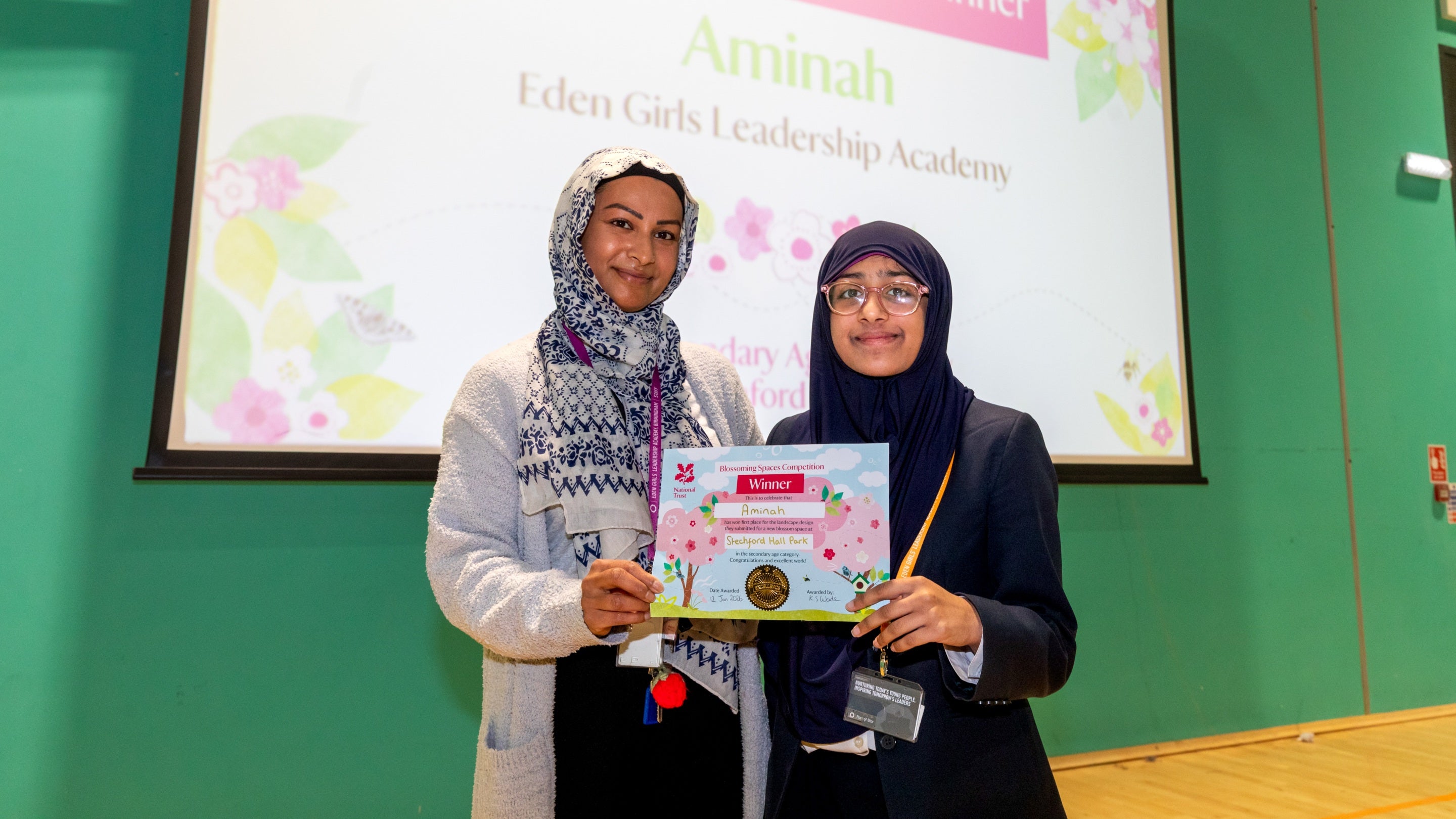 A woman and a child both in headscarves holding a certificate between them. behind them a screen says Aminah Eden Girls leadership Academy