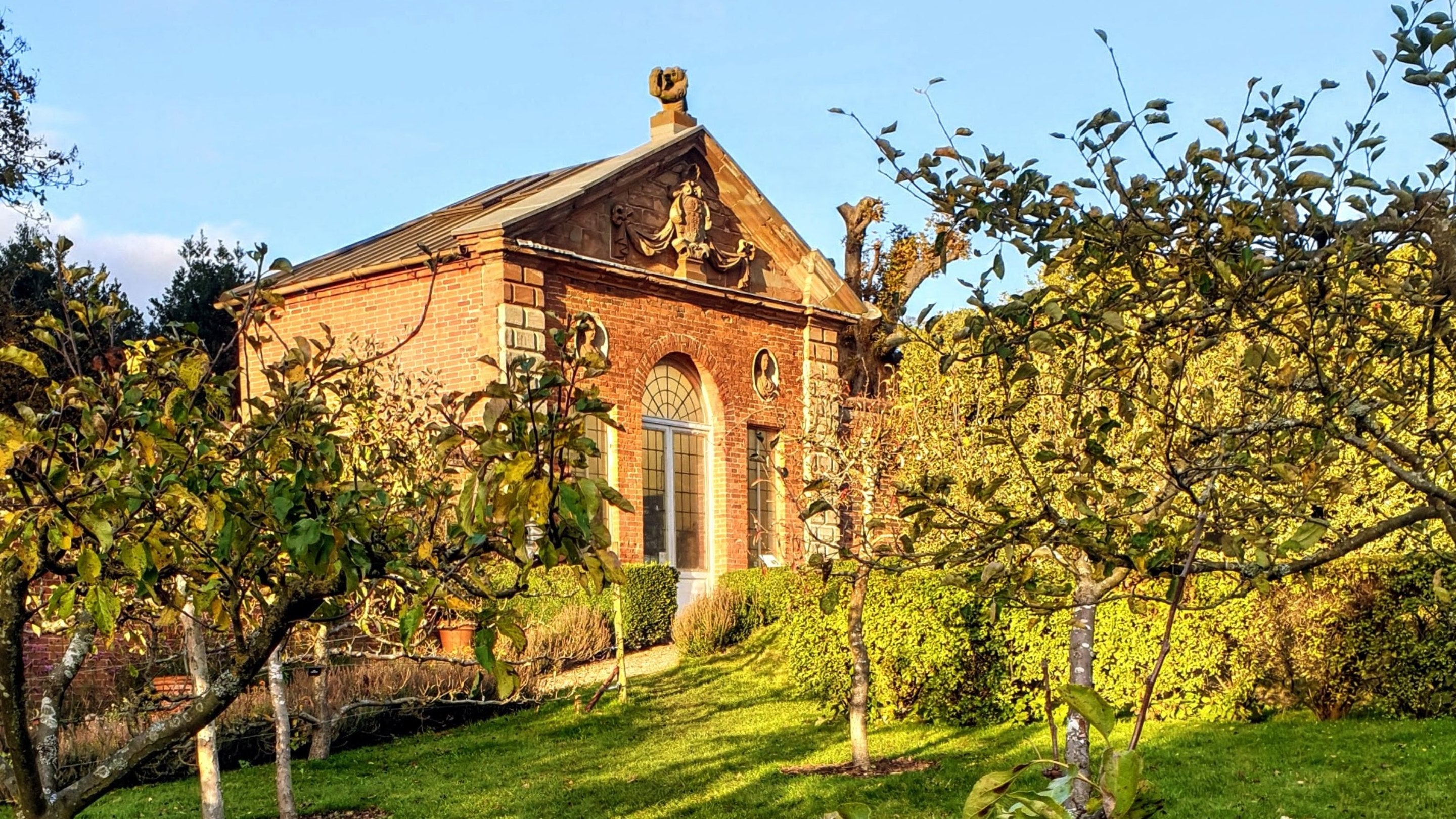 A decorative, red bricked building with a peaked roof and white window frames is glimpsed between autumnal apple trees, in the low autumn sunlight which makes everything glow.
