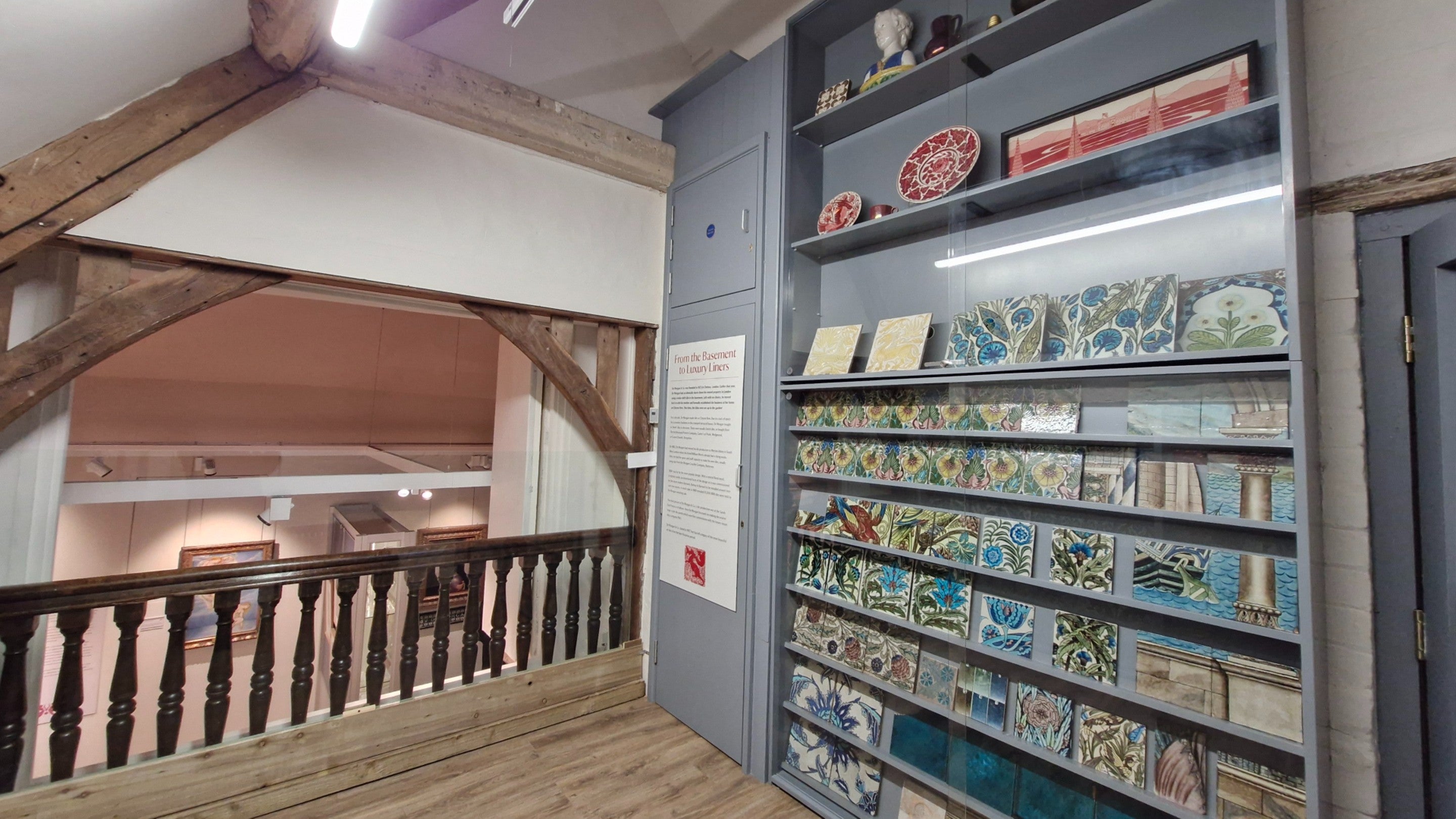 A view of a ceiling-height cabinet with a glass front which contains colourful tiles by William De Morgan. The tiles show images of bird, plants and architecture.