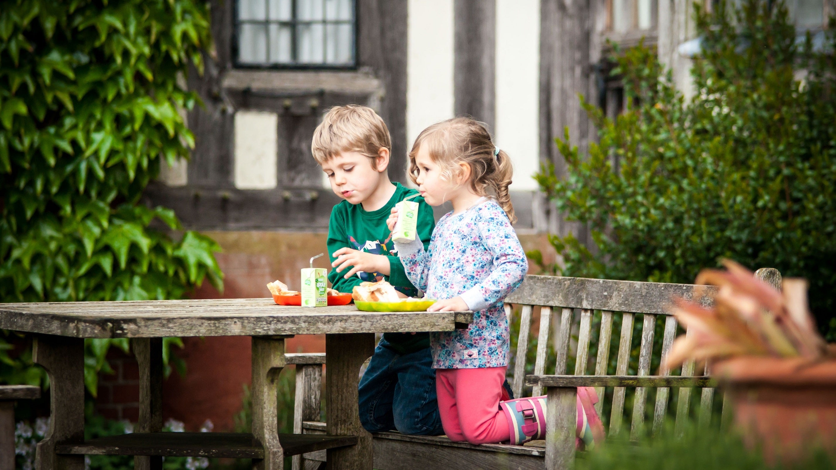 Two children are sitting at a table on the South Terrace at Wightwick Manor. They have lunchboxes and are eating sandwiches and drinking from cartons through a straw.