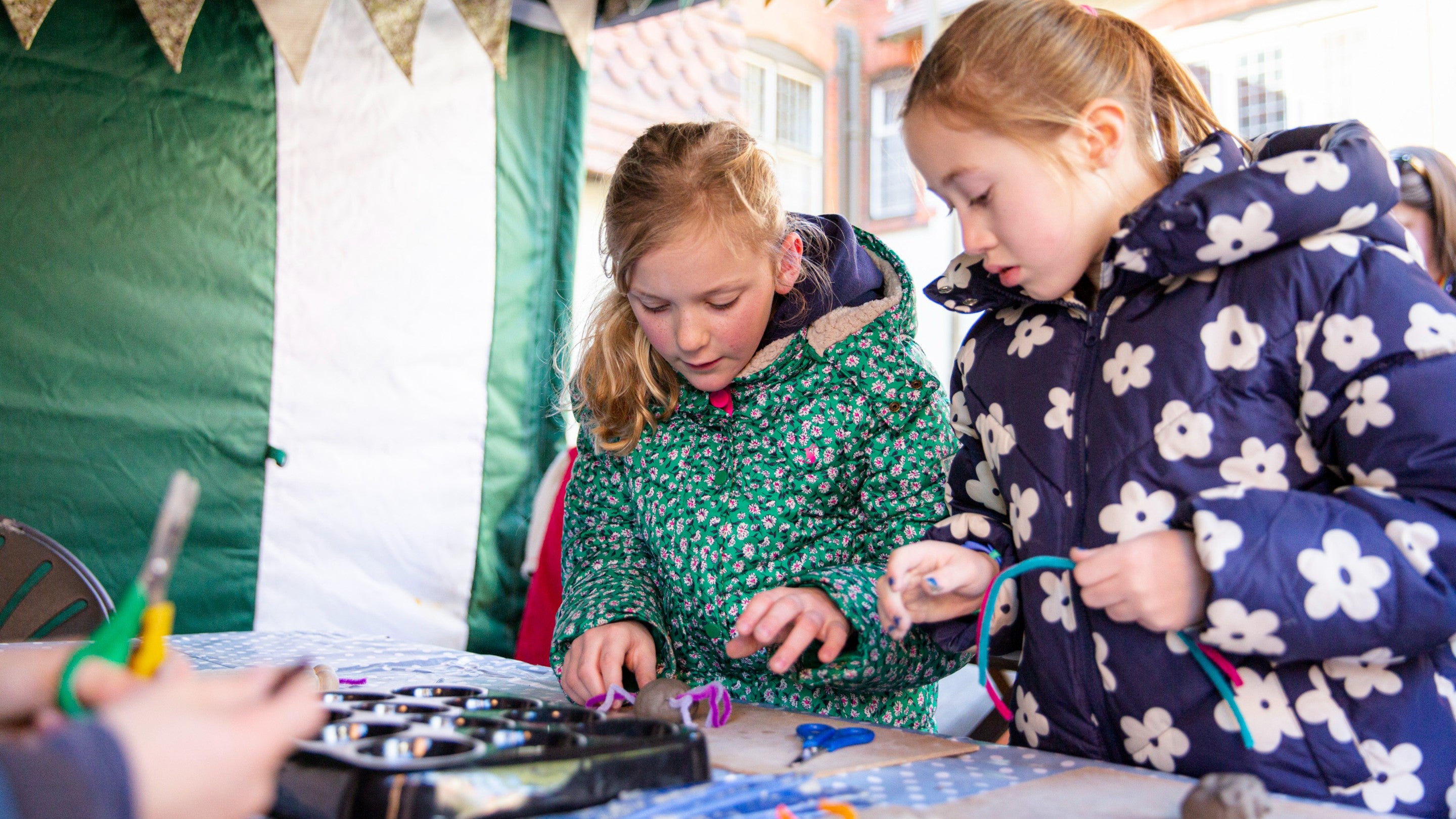 Two girls are working at a table under a gazebo, one is moulding clay whilst the other holds a pipe cleaner.