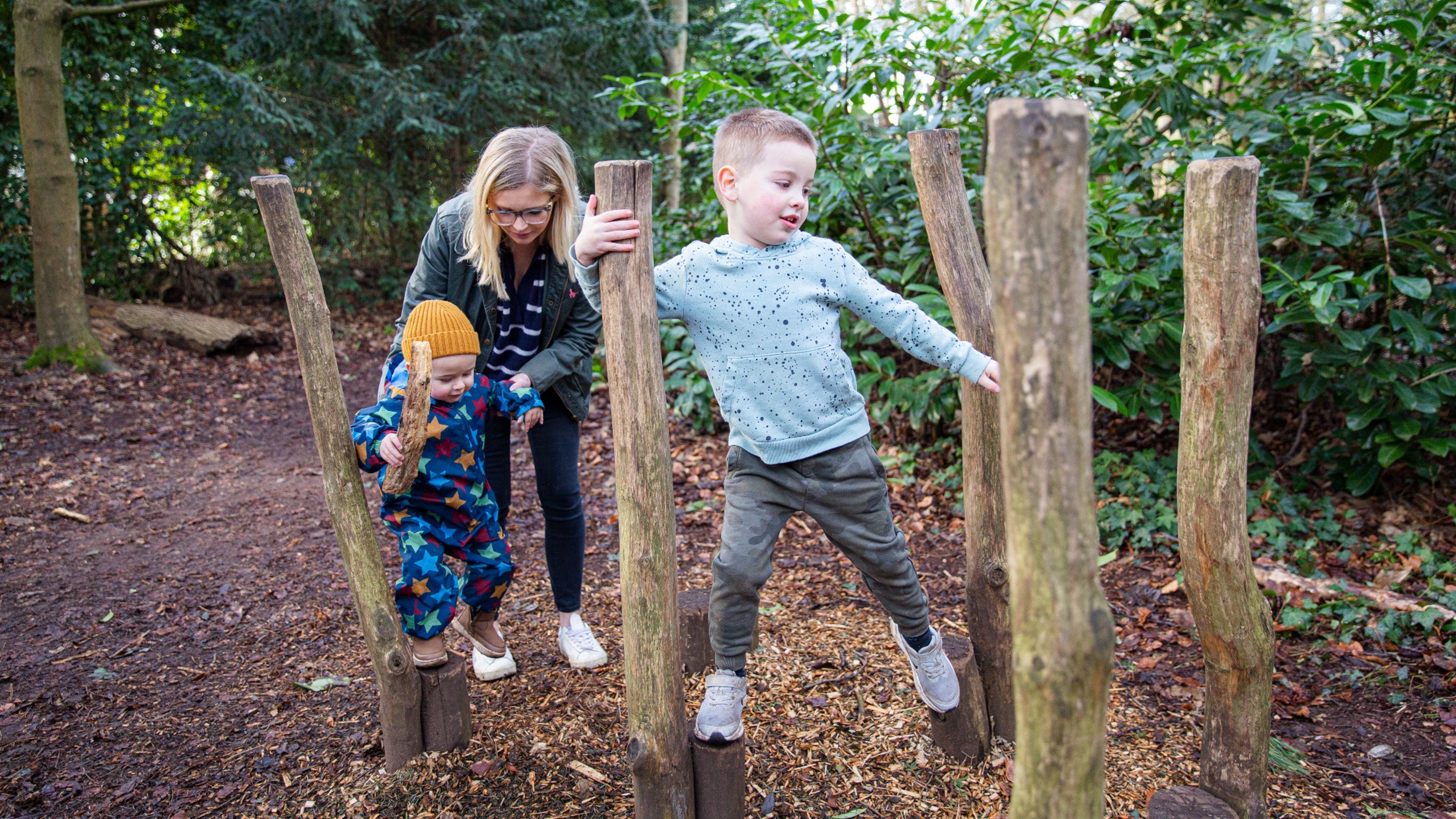 Two young children are walking along wooden raised stilts in the woodland play at Wightwick Manor. The younger child at the back is being helped by an adult.
