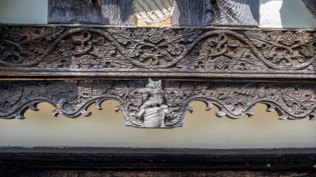A close view of an intricately carved oak beam which has silvered down with age. Carved into the centre is a figure of a fox or wolf type creature holding what seems to be a shield. It is surrounded by twisted leaves and other natural shapes.