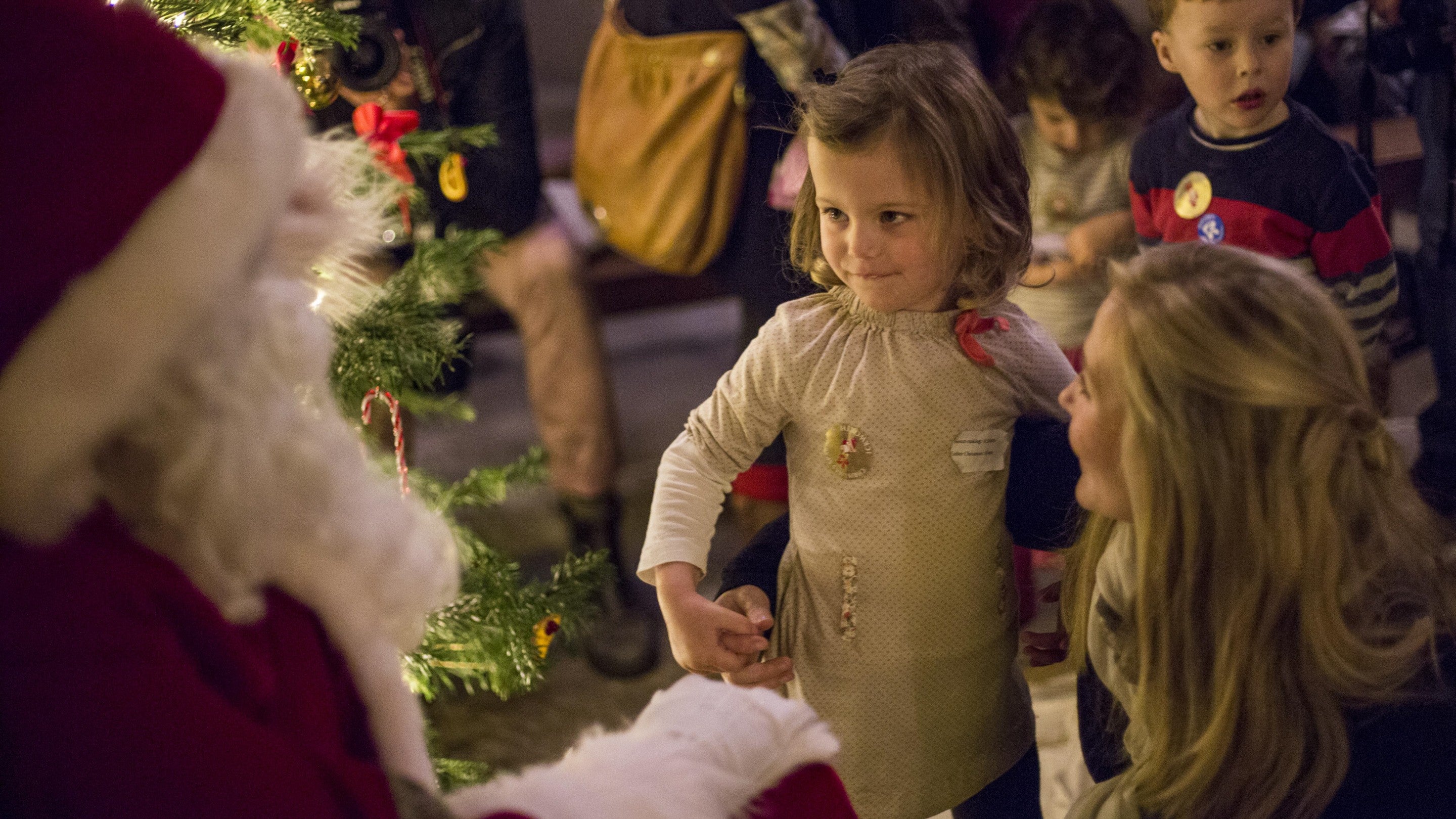 Father Christmas in his red suit, is giving a gift to a child. There is a Christmas tree in the background and other children waiting.