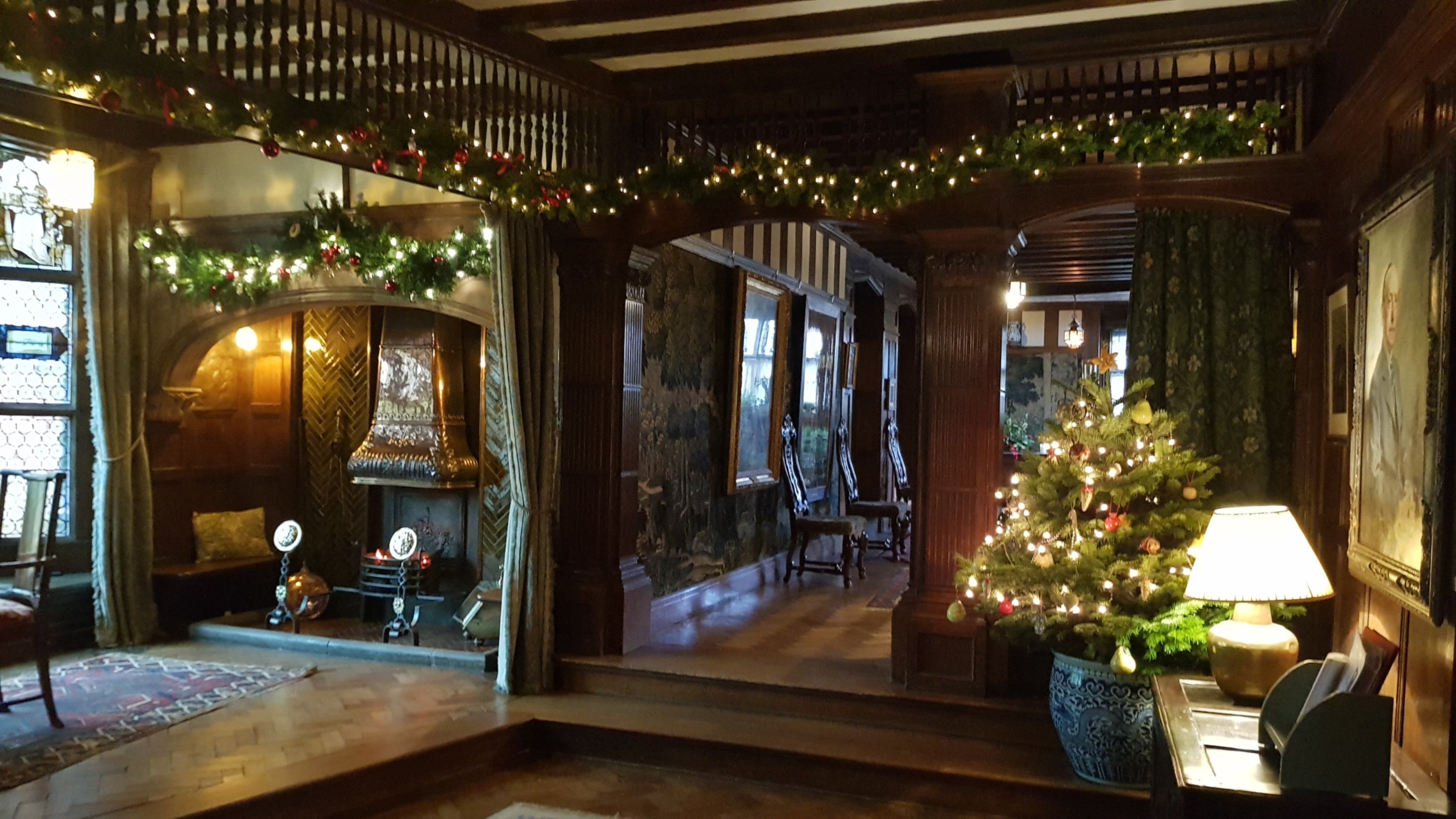 A view of the Lower Hall at Wightwick Manor. There are lights around the tops of the fireplace and walls and there is a Christmas tree to the right of the steps.