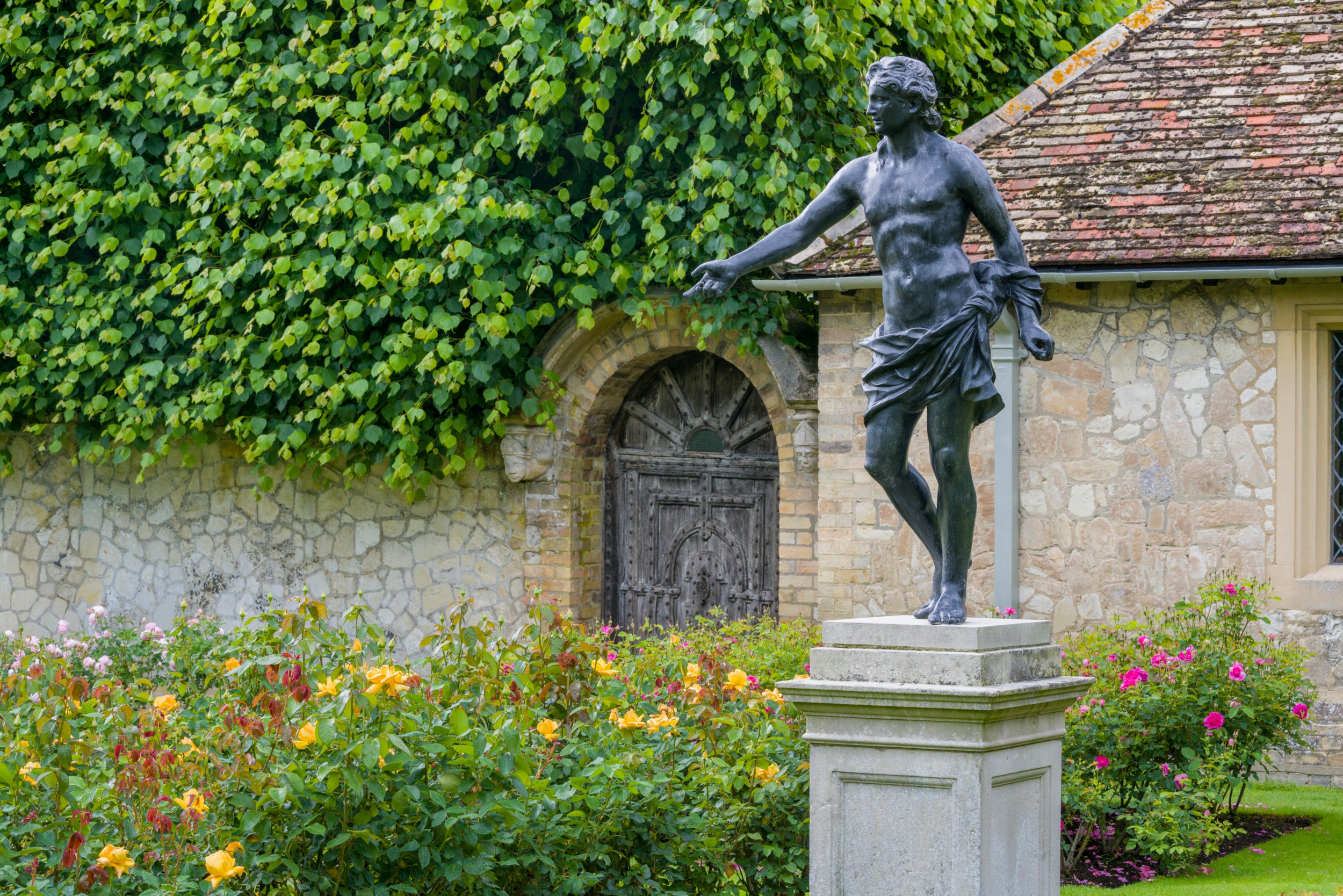 Apollo sculpture in the Rose Garden at Anglesey Abbey. The statue is on a raised stand, arm outstretched and surrounded by yellow roses.