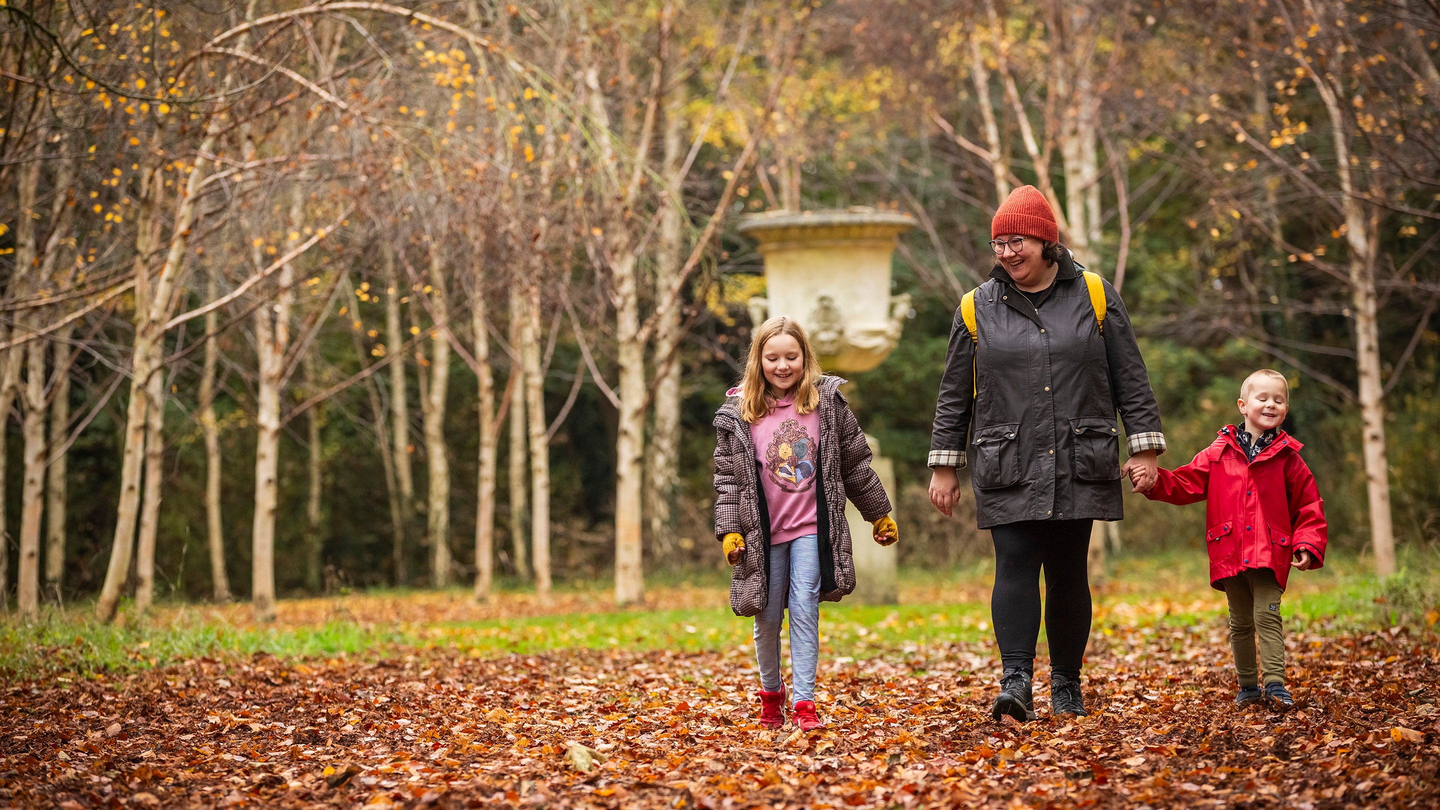 A woman and two children, dressed in warm coats and bobble hats, walking through fallen leaves with autumn trees and a garden statue in the background