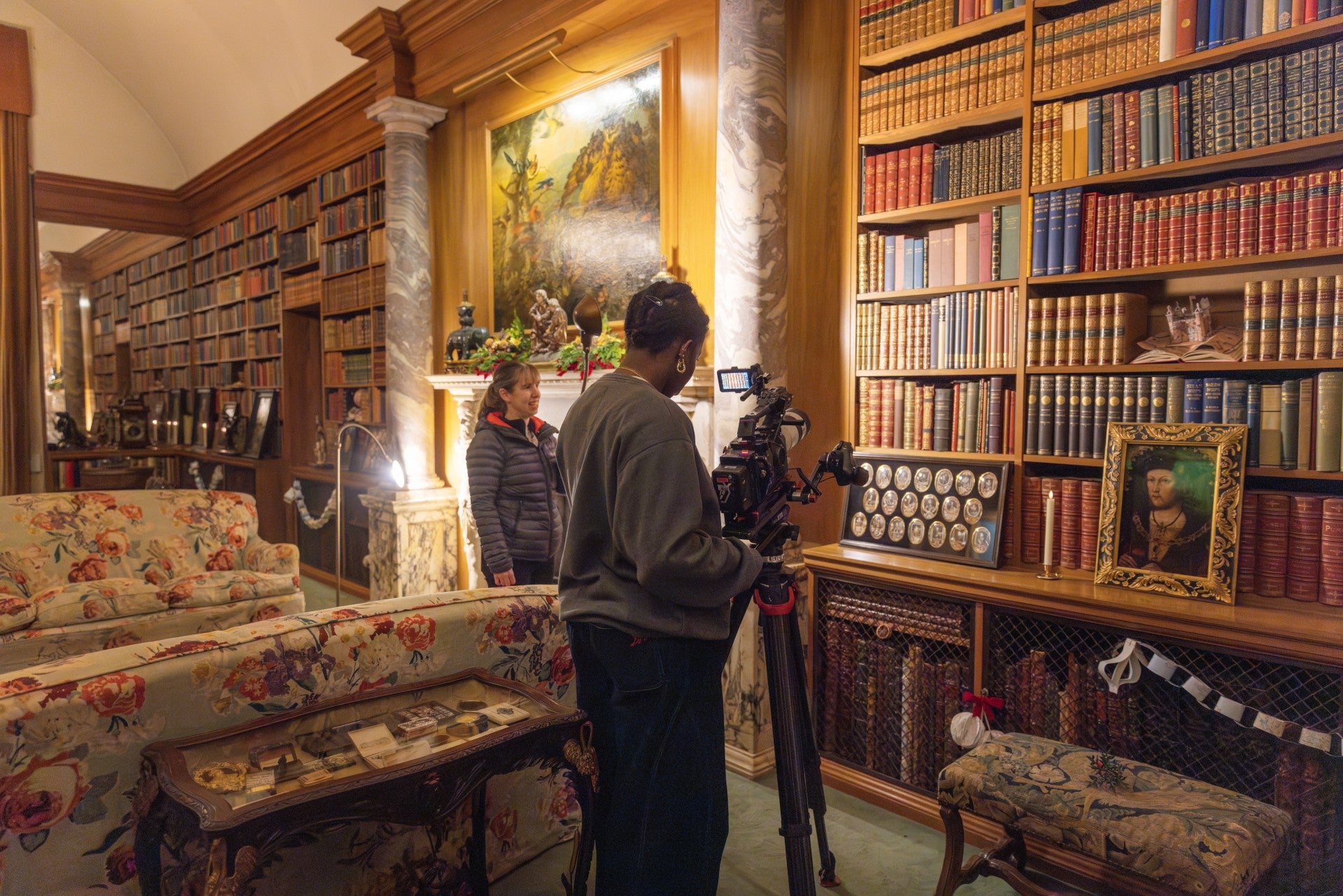 A camera lady is filming decorations in front of the bookshelves in the Anglesey Abbey library. A member of staff is assisting..