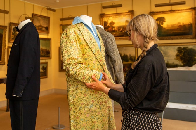 A lady dressed in black and standing at the right of the photo is making adjustments to Lord Fairhaven's gold silk dressing gown on a mannequin. There is another black suit and lots of paintings in the background.