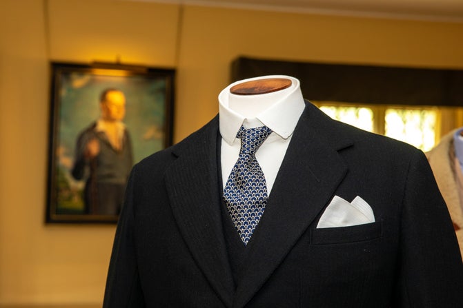 A close up of the top half of Lord Fairhaven's black morning coat, his portrait hanging on the wall behind.