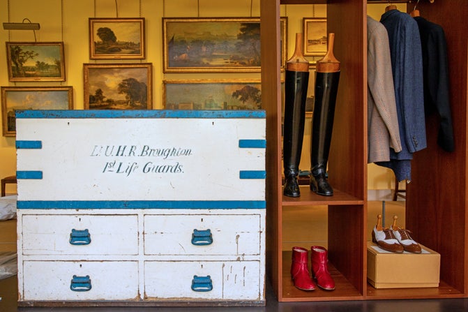 Lord Fairhaven's white and blue travelling trunk, with the writing 'Lt U.H.R Broughton, 1st Life Guards' inscribed on it. Long black boots and small red leather boots alongside suits hanging up in a wardrobe next to the trunk.