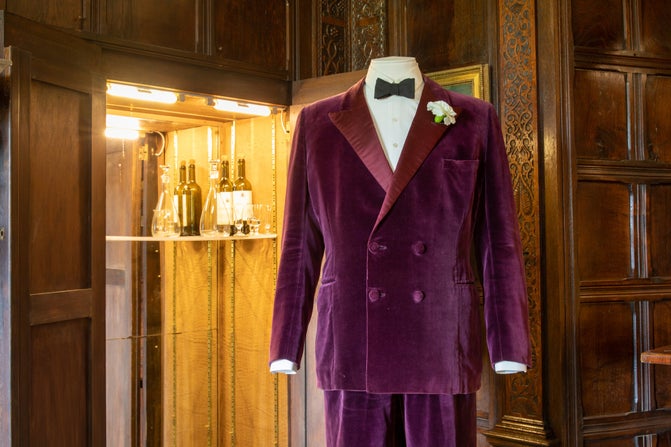Top half of Lord Fairhaven's wine-coloured velvet smoking suit on the right and white carnation in the pocket, with a cupboard open behind it and to the left, with a light on and wine bottles on a shelf. Brown panels in room.