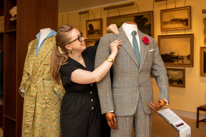 Roisin Semple checks the lapel of Lord Fairhaven's grey tweed jacket in the Tailoring an Image exhibition. She is standing between two mannequins, the other has a golden silk dressing gown on it.