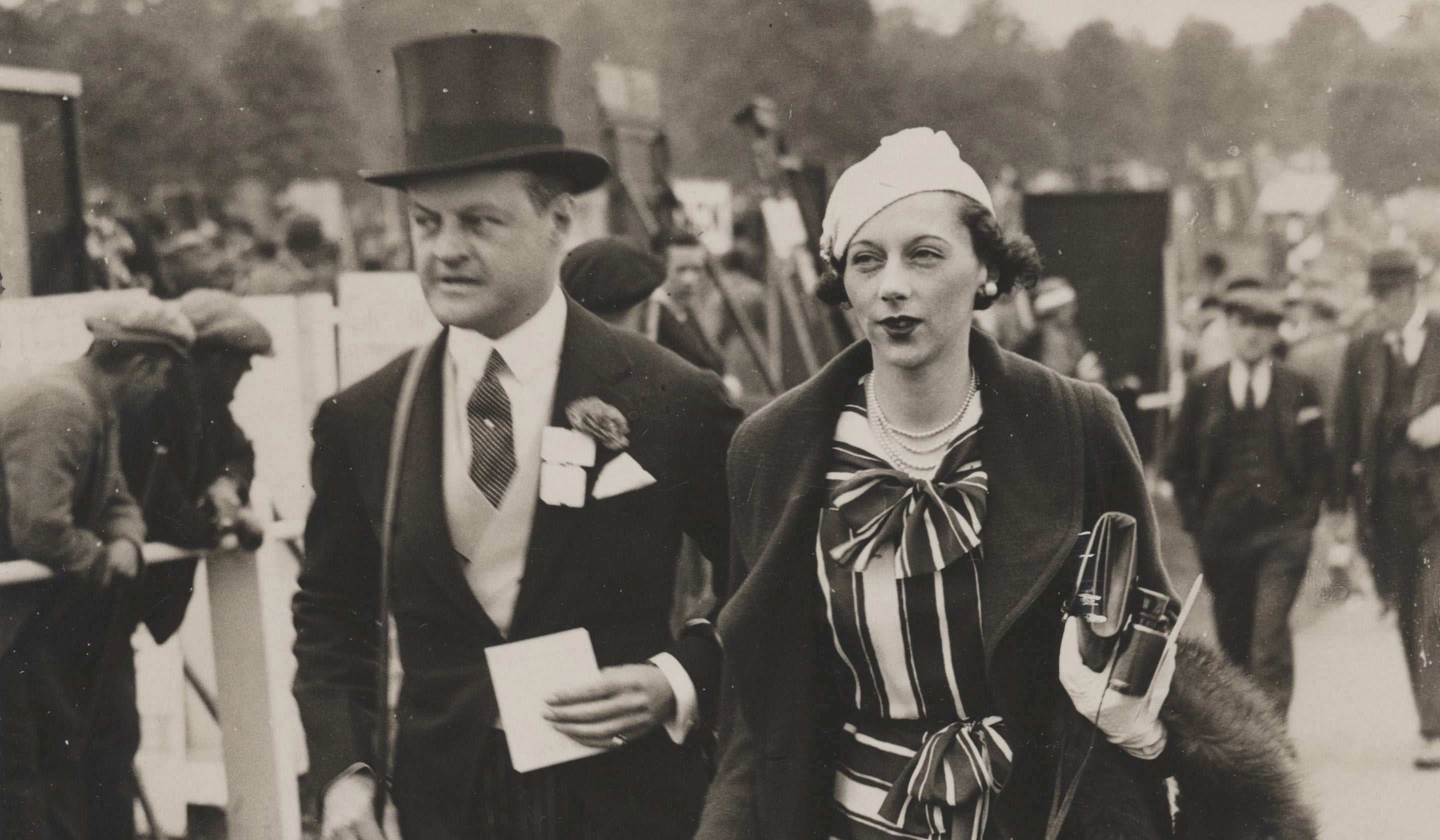 A black and white photo showing a gentleman in top hat and tail coat escorting a lady at a horse racing event