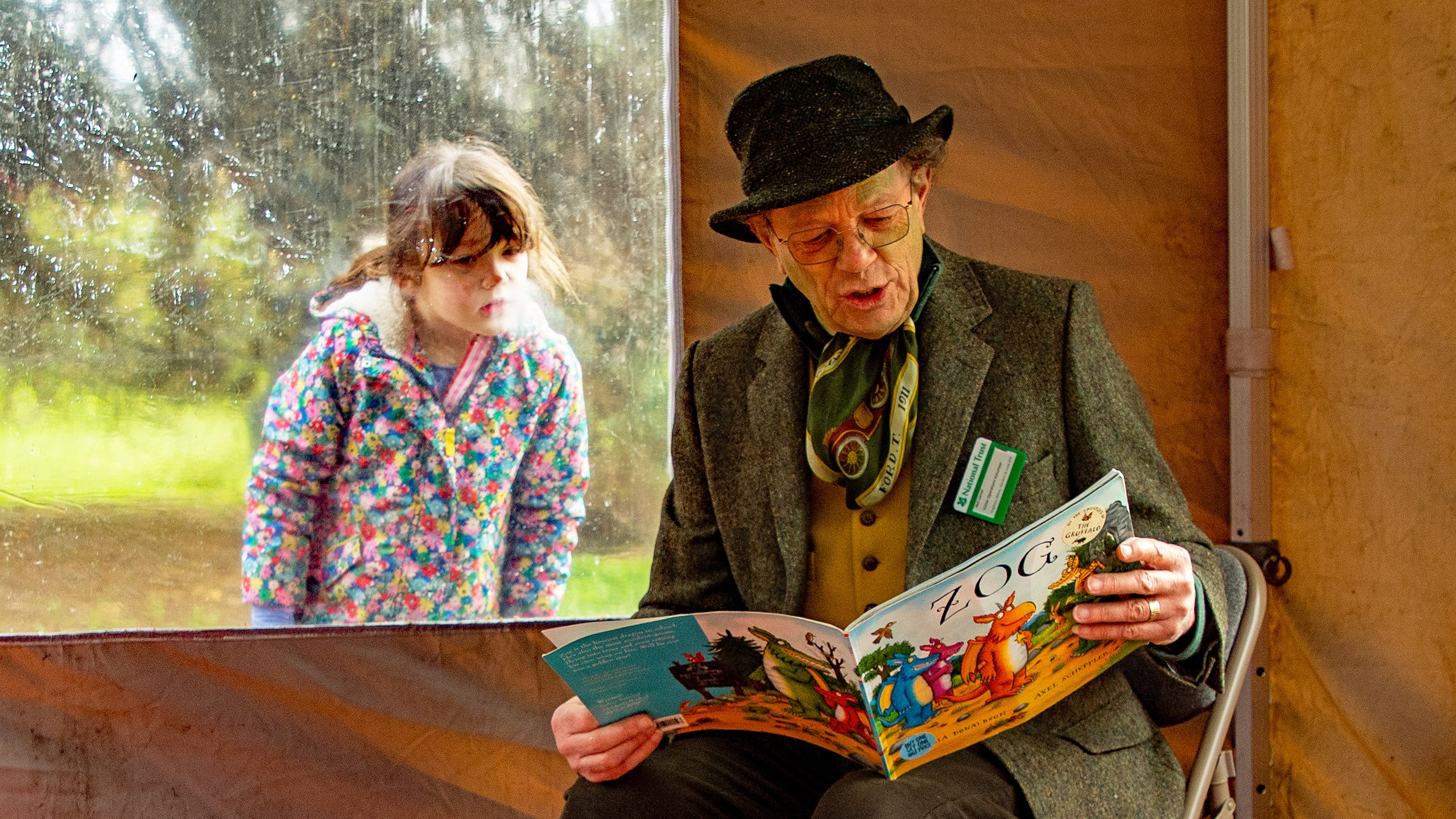 A man wearing a bowler hand and waistcoat reads the picture book "Zog" while sitting in a tent. A young girl peers in through the tent window to watch him.