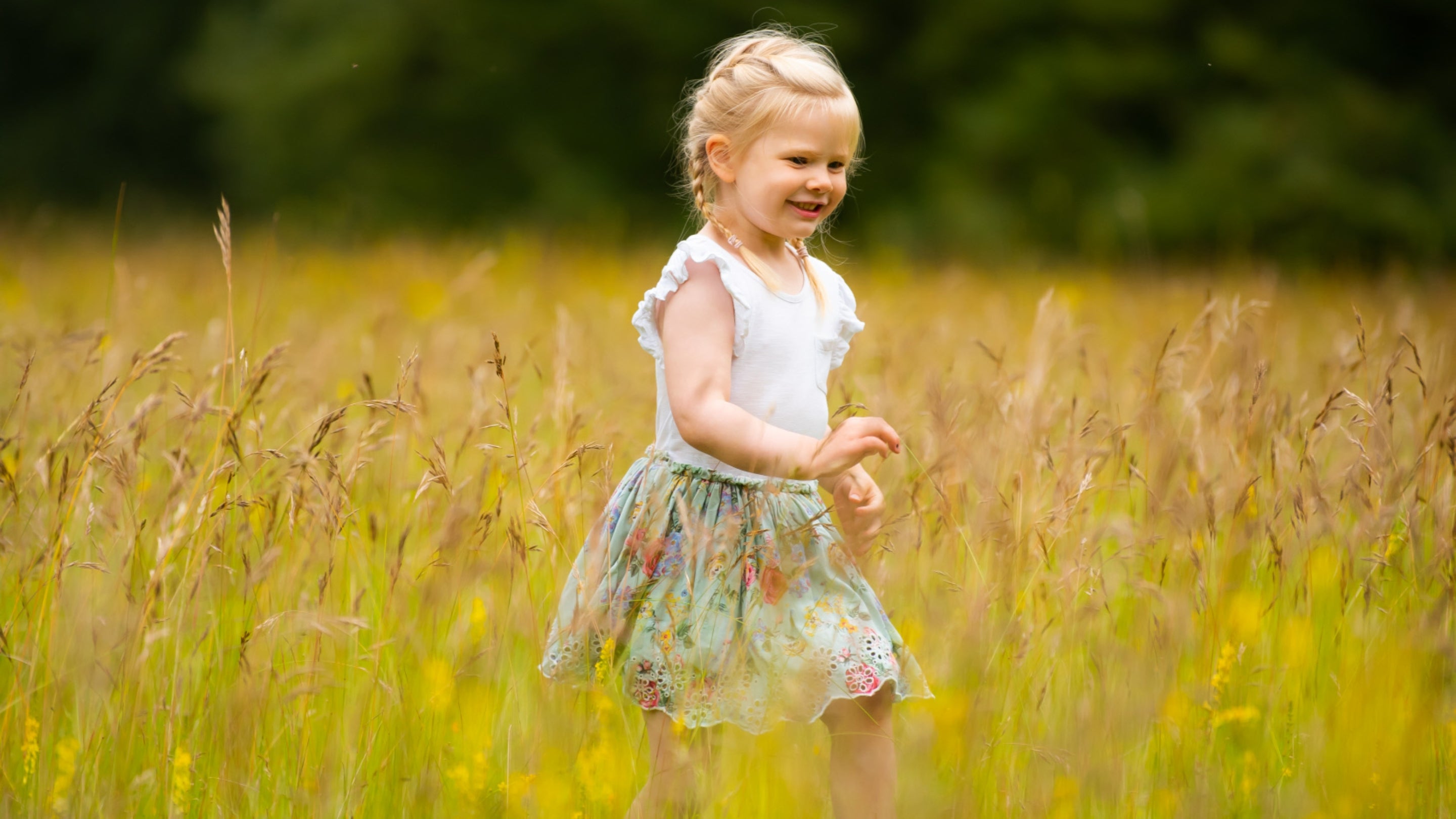 A girl plays in long grass in a meadow with trees in the background.