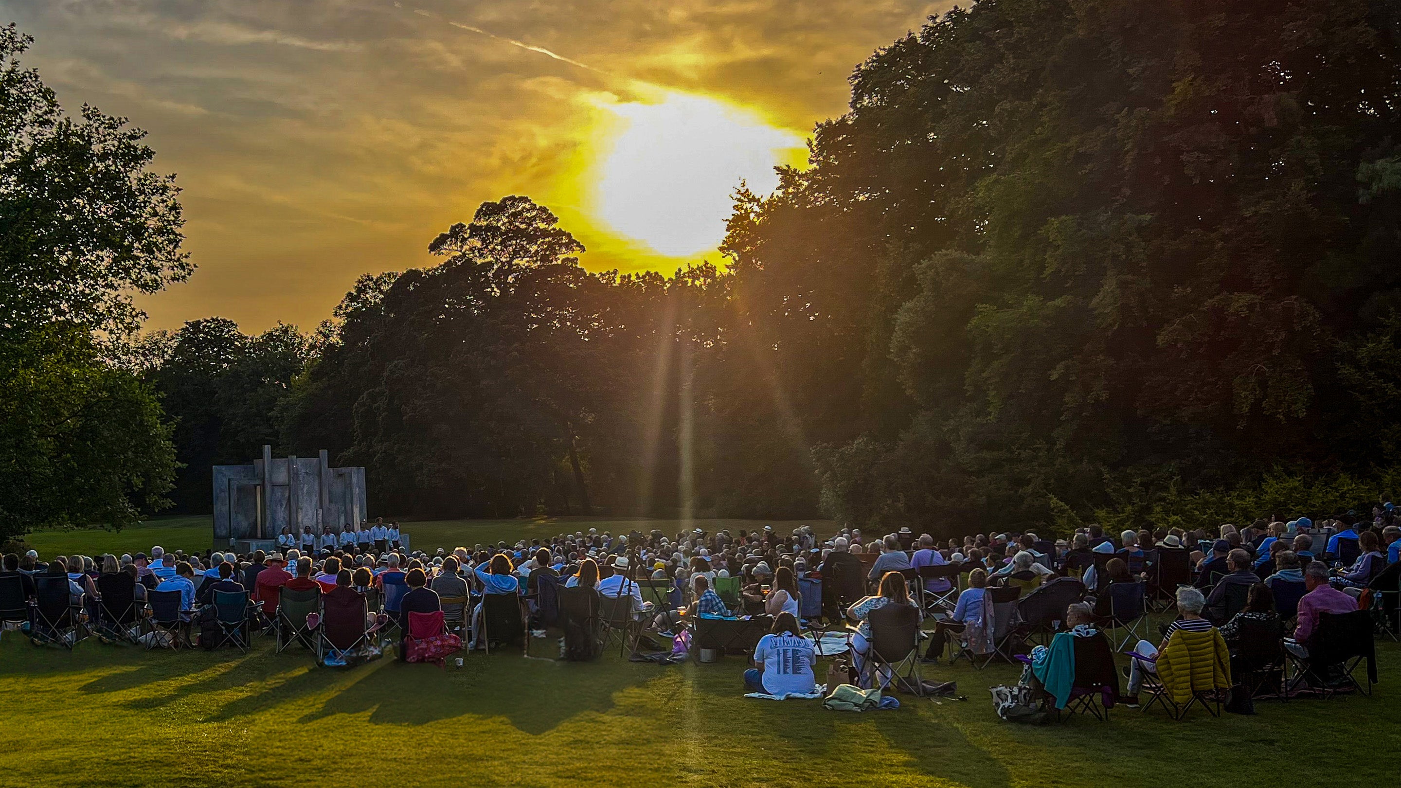 An audience watching an outdoor theatre performance in a parkland at sunset